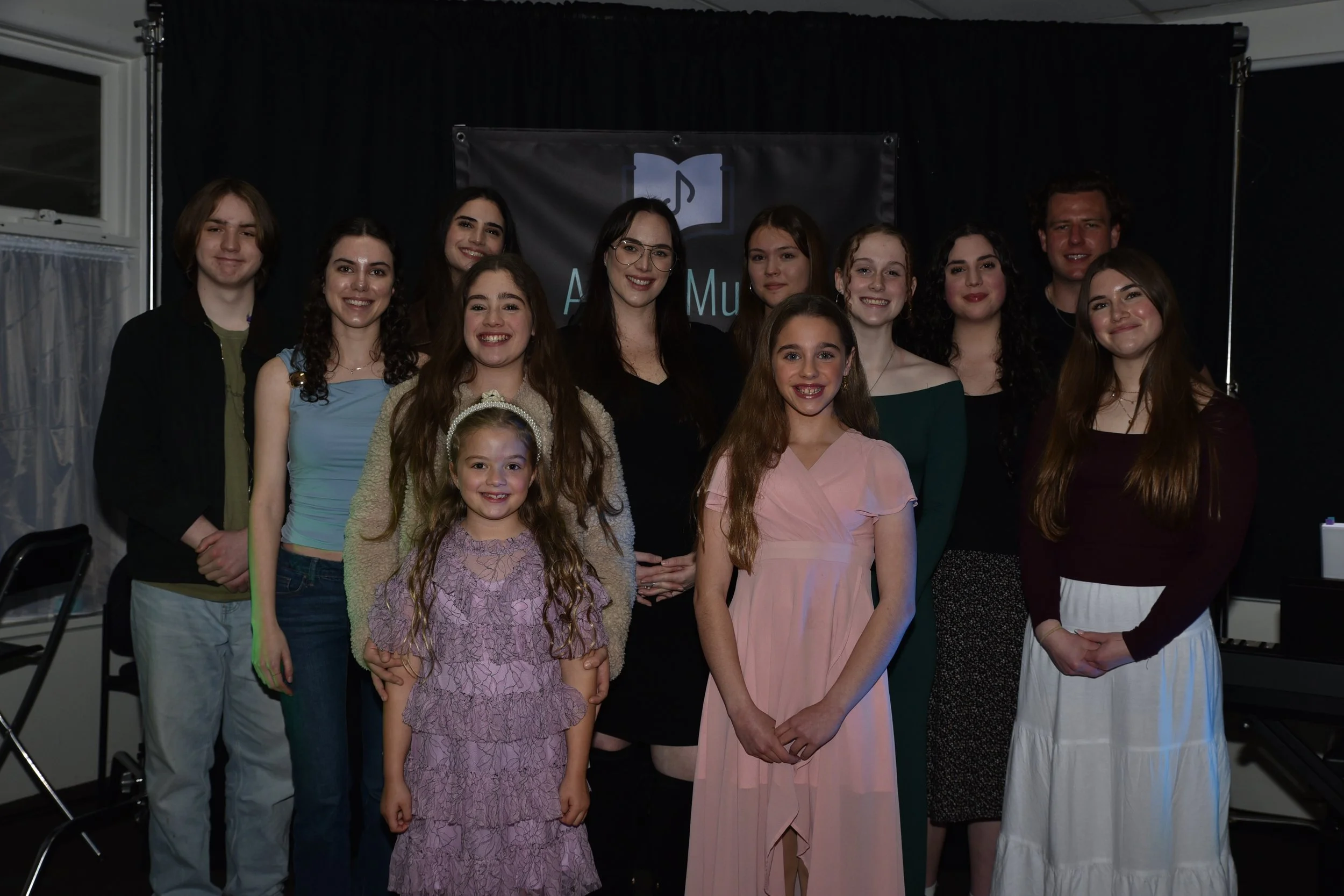 A group of fourteen people, mostly young women and two men, standing together indoors in front of a black curtain, smiling at the camera.