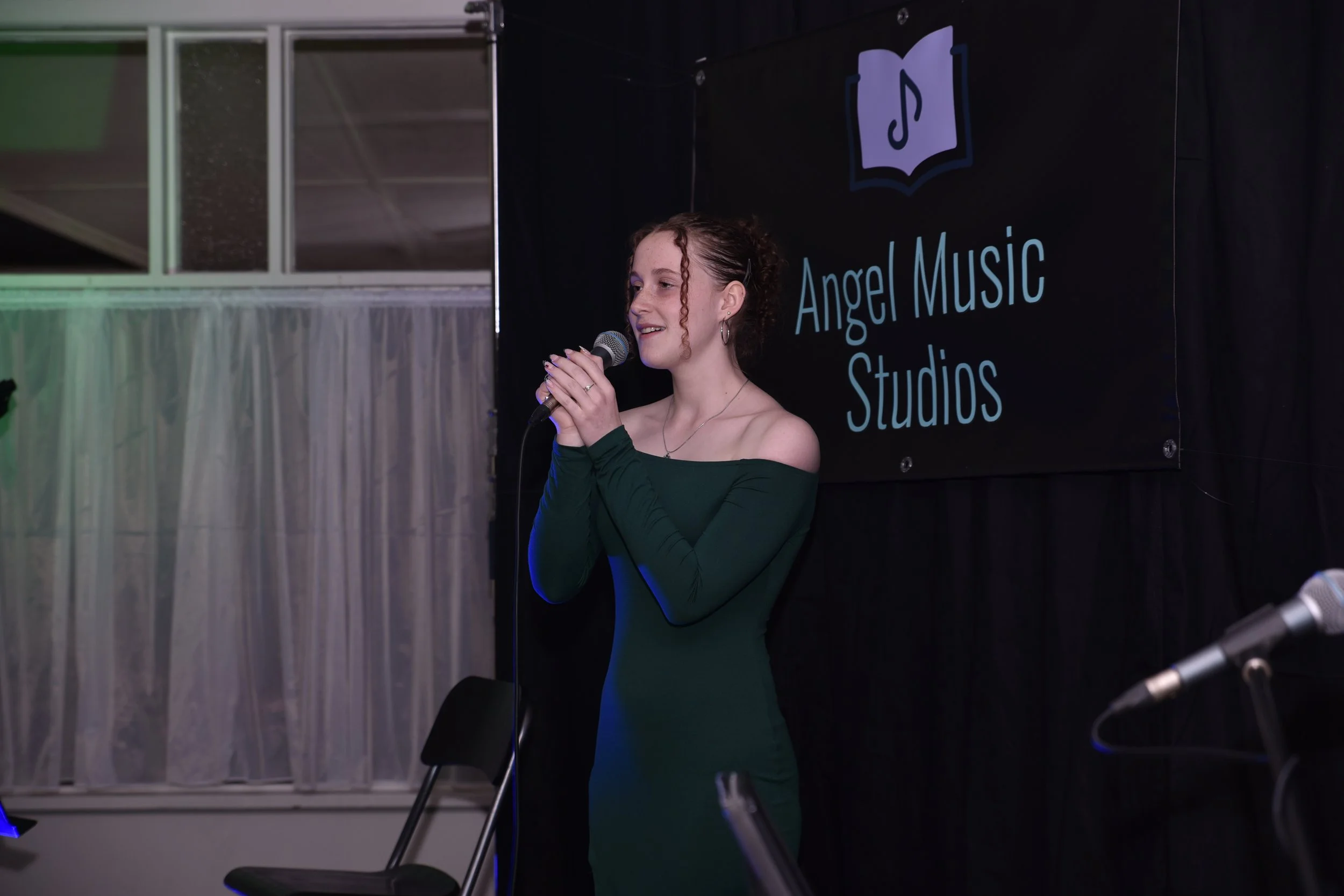 A young woman with curly brown hair, wearing a dark green off-shoulder dress, holding a microphone and singing on stage at Angel Music Studios.