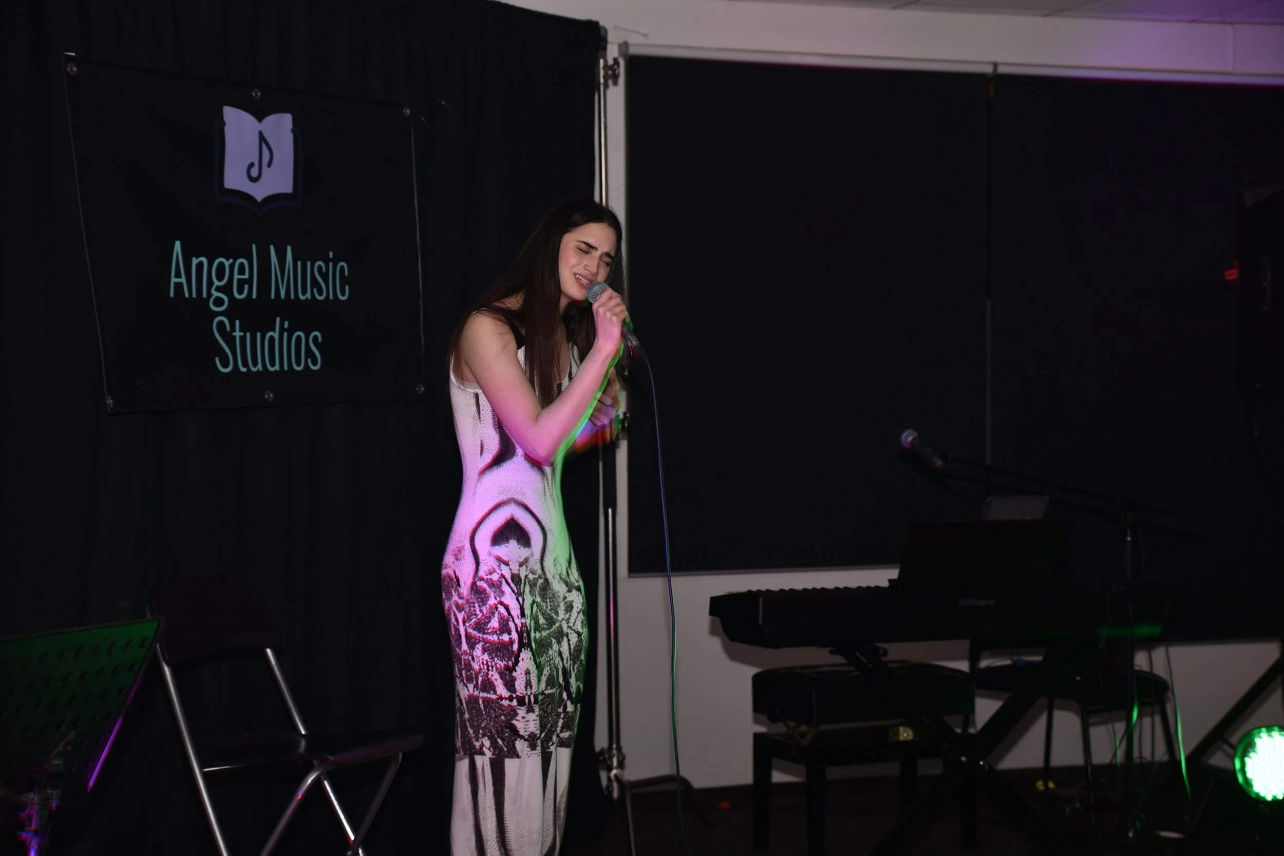 A woman singing passionately into a microphone on stage at Angel Music Studios with musical equipment nearby.