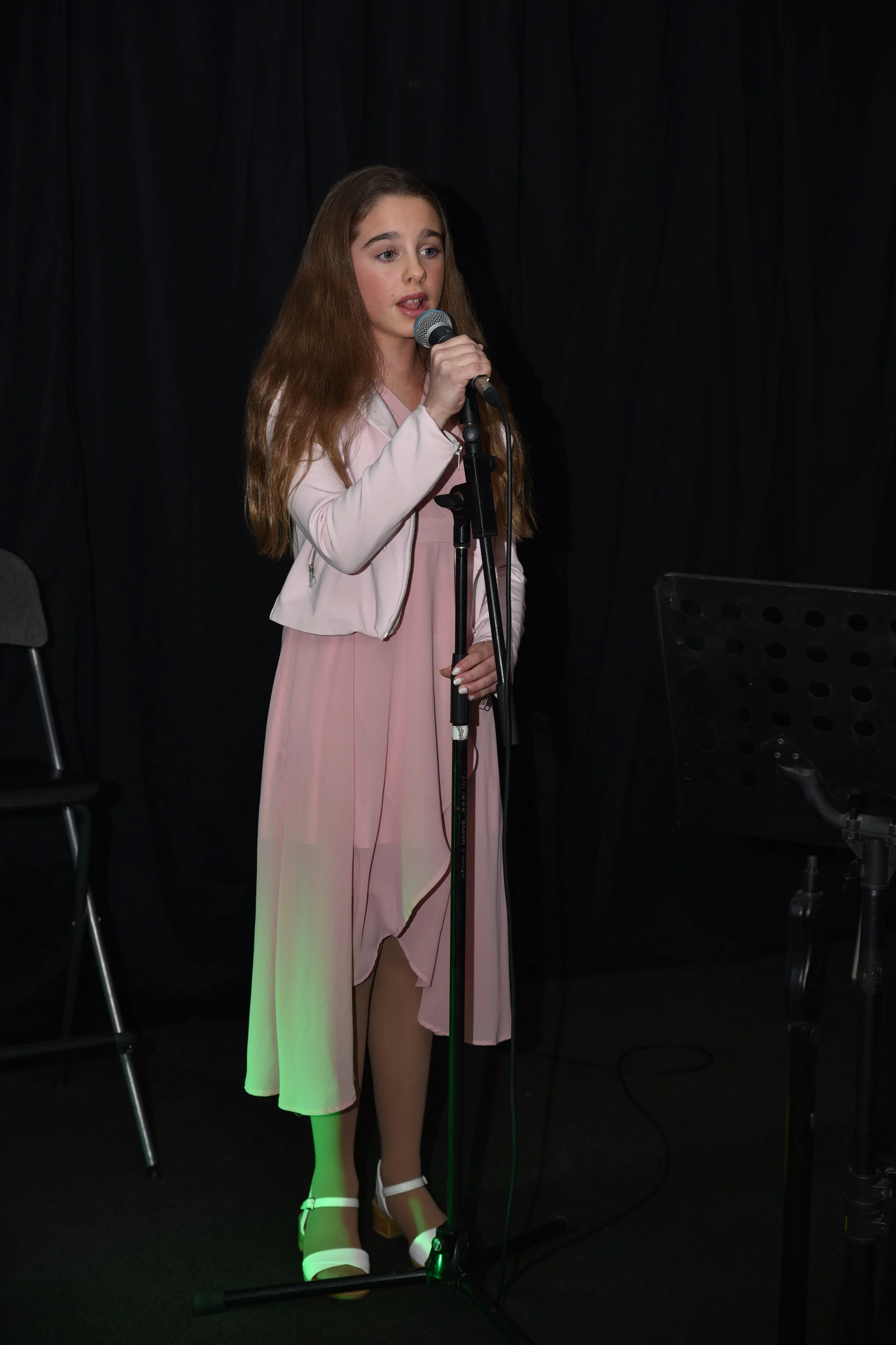 Young girl with long brown hair singing into a microphone on a stage with black curtains, dressed in a pink dress with a matching light pink jacket and white shoes.