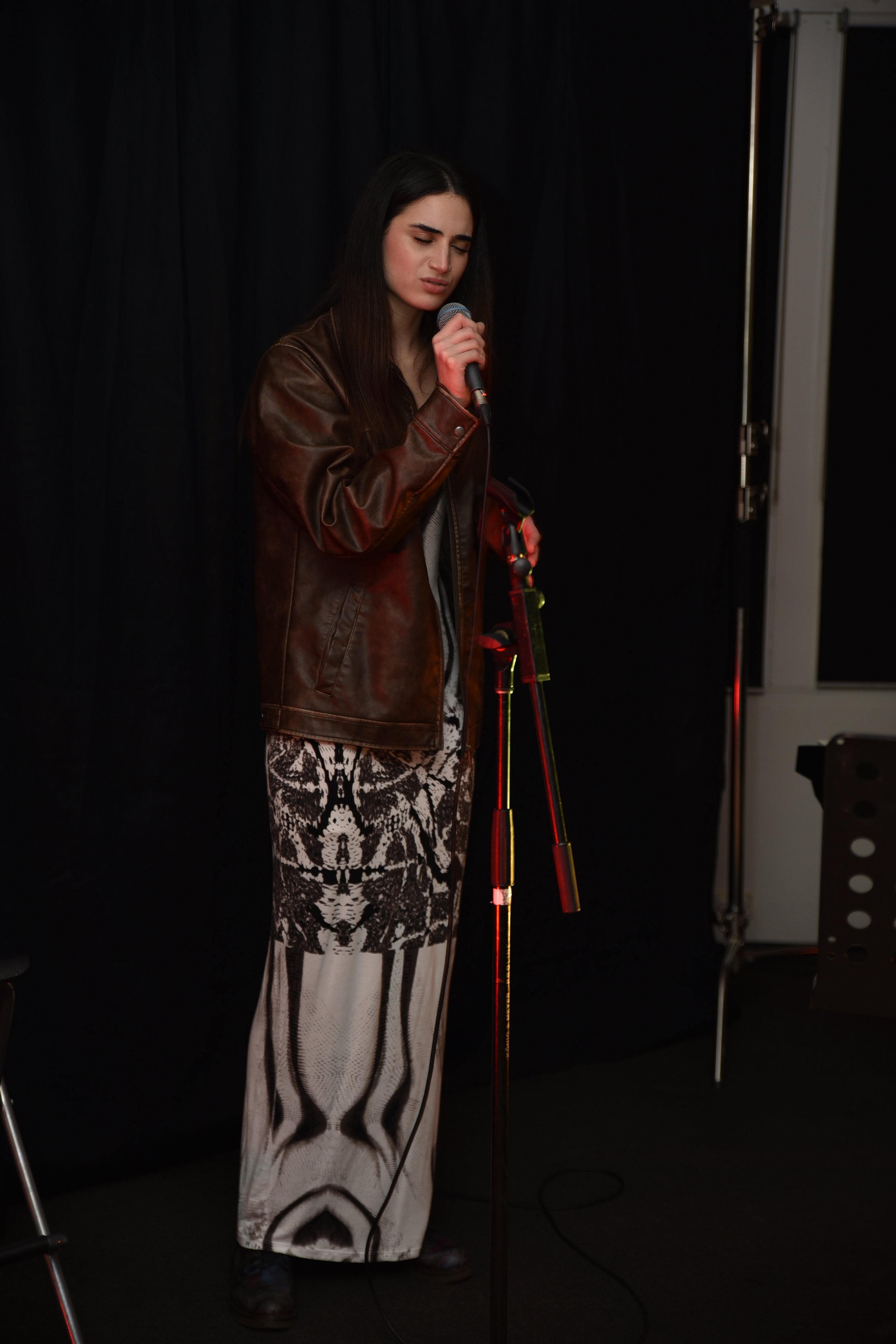 A woman with dark hair is standing in front of a black curtain, holding a microphone and singing or speaking. She is wearing a brown leather jacket, a long patterned skirt, and standing in a dimly lit setting with stage equipment visible.