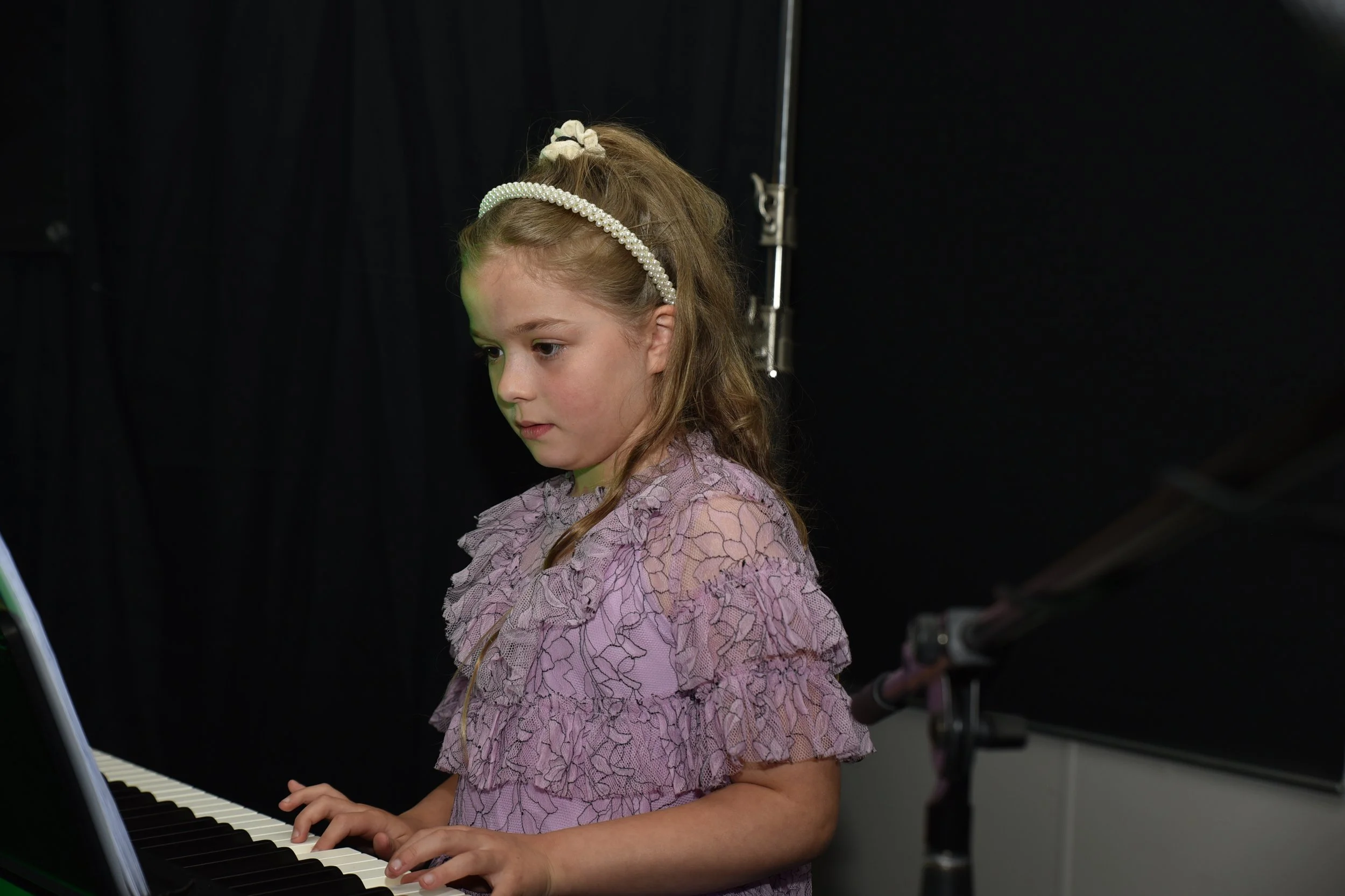 A young girl with a pearl headband playing the piano, wearing a pink lace dress with floral patterns, in a room with a black backdrop.