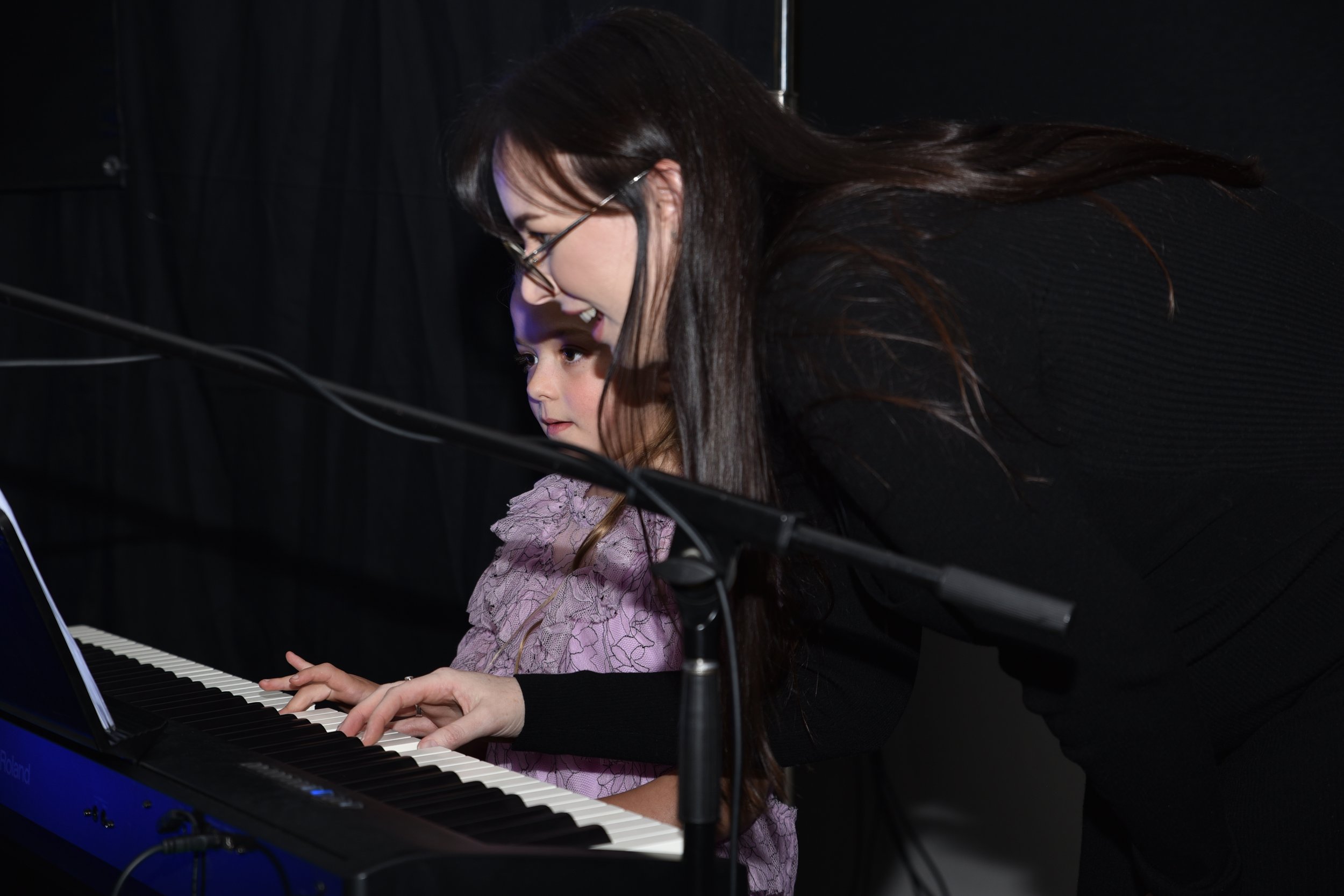 A woman and a young girl playing a piano together in a dark room with a black background.
