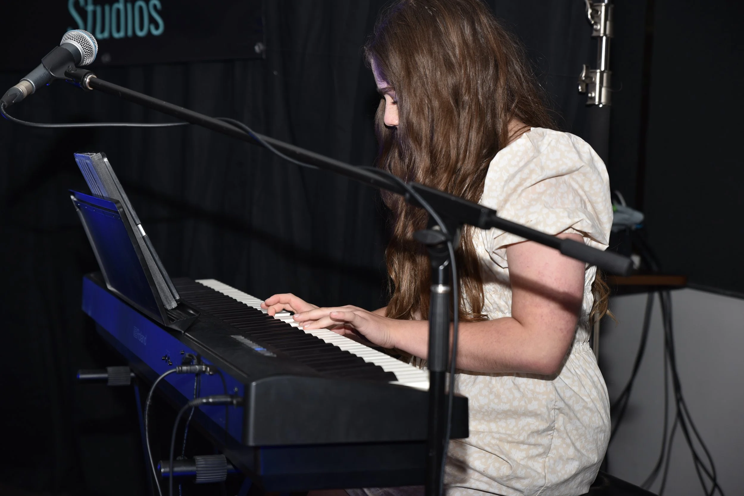 A young woman with long, wavy brown hair wearing a light-colored floral dress, playing a blue digital piano in a recording studio with a black curtain backdrop.