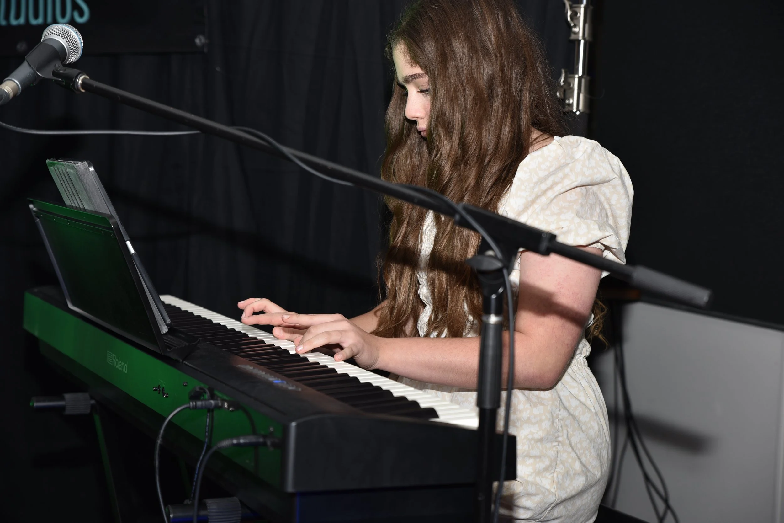 A young woman with long brown hair playing a green Roland digital piano on stage with a black backdrop.