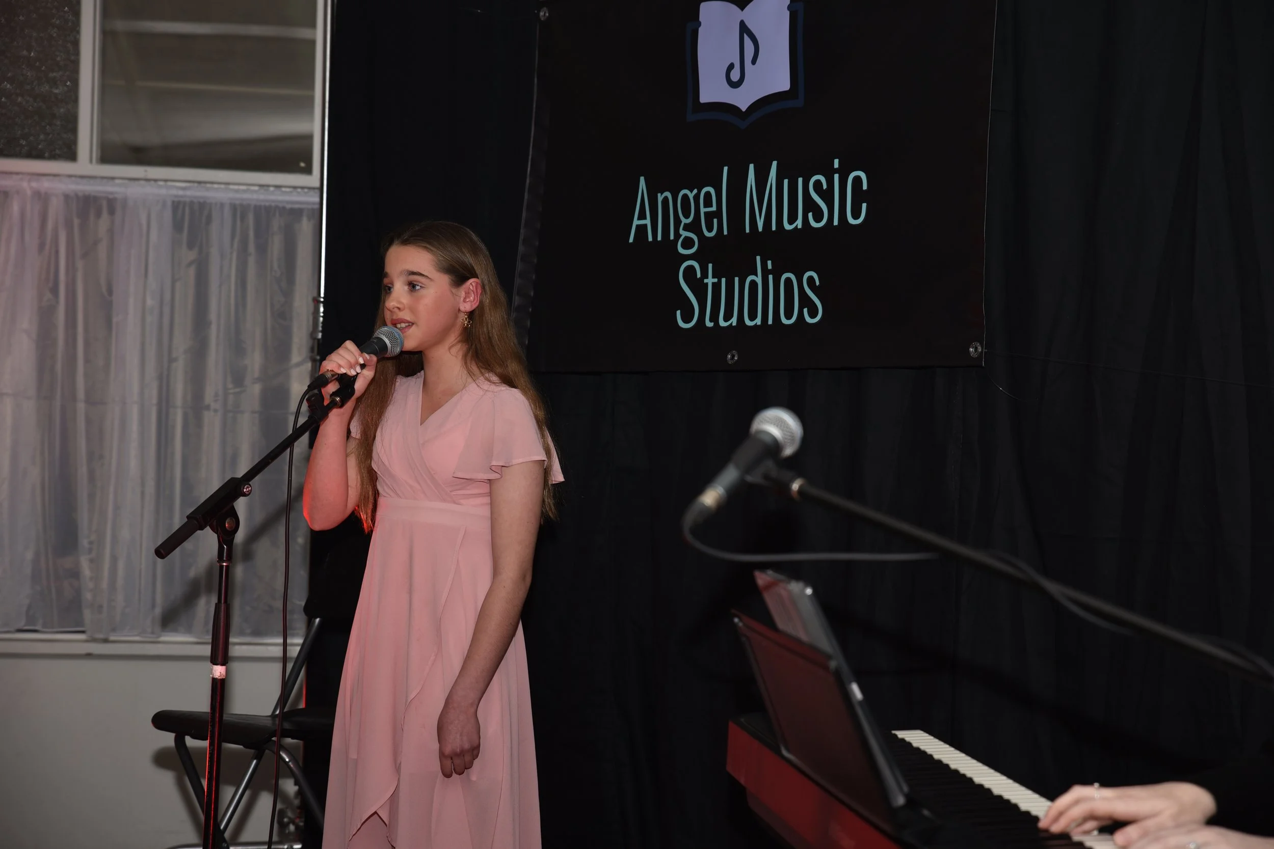 Young girl in pink dress singing into a microphone on stage, with a sign in the background reading 'Angel Music Studios' and a person playing a piano in the foreground.