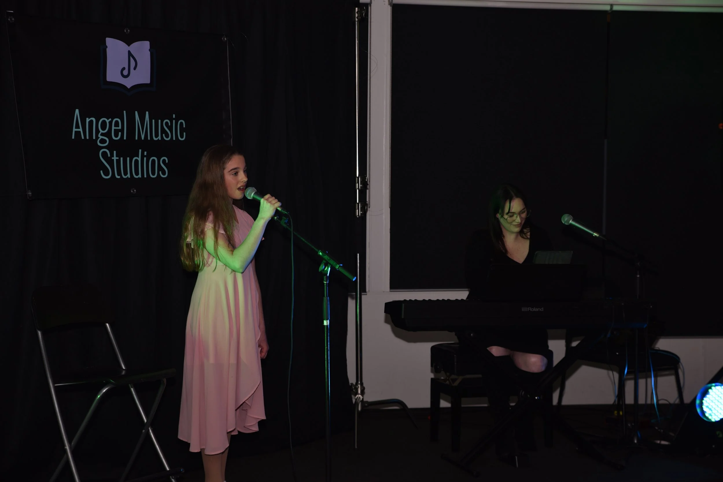 Young girl singing into a microphone on stage at Angel Music Studios, with a woman playing a keyboard beside her.