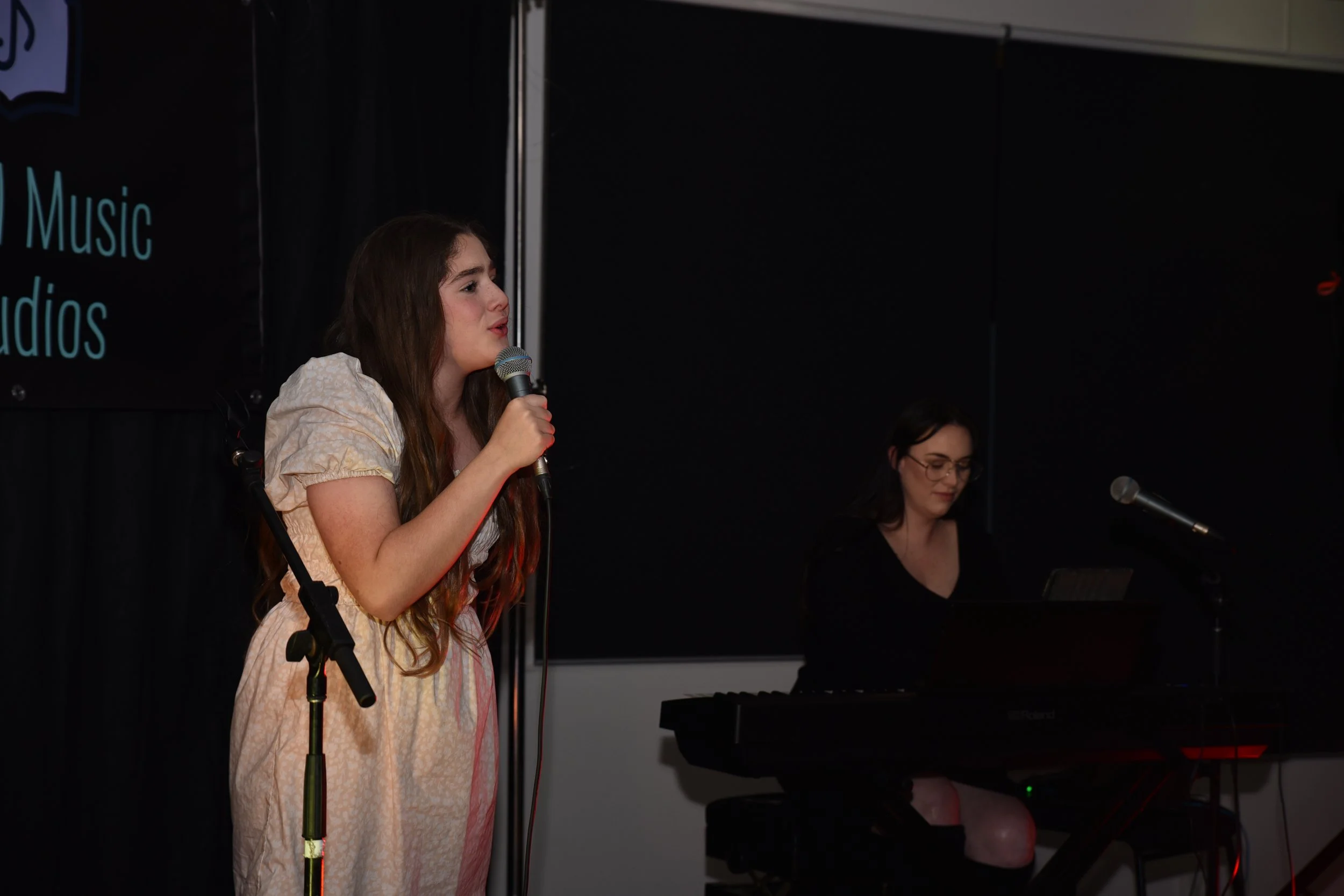 A young woman with long wavy brown hair singing into a microphone on a stage, wearing a light-colored dress. Next to her, another woman with glasses is playing a keyboard. The background is black with a partially visible sign on the left that include