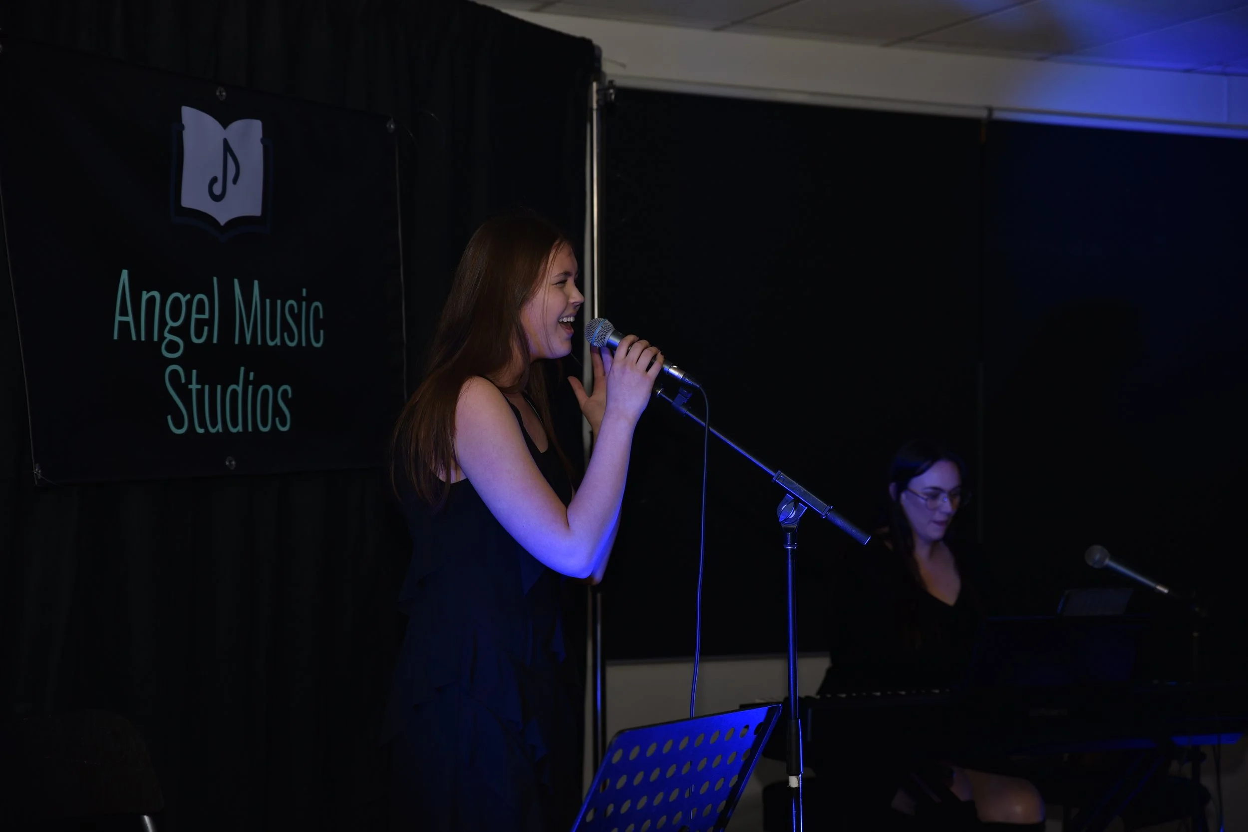 A young woman with long reddish-brown hair singing into a microphone on stage at Angel Music Studios. She is smiling and wearing a black sleeveless top. There is a black backdrop with the Angel Music Studios logo and name, and another person playing 