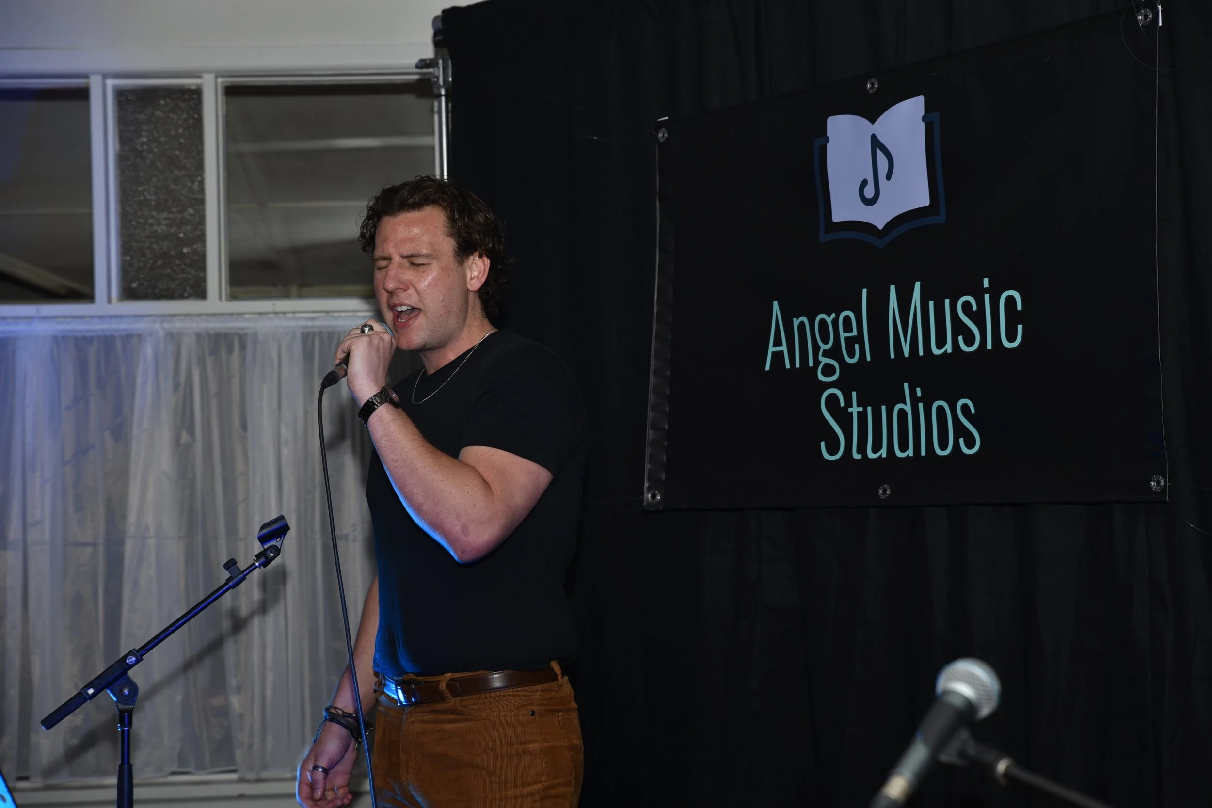 A man singing into a microphone at an event with a sign behind him reading 'Angel Music Studios'.