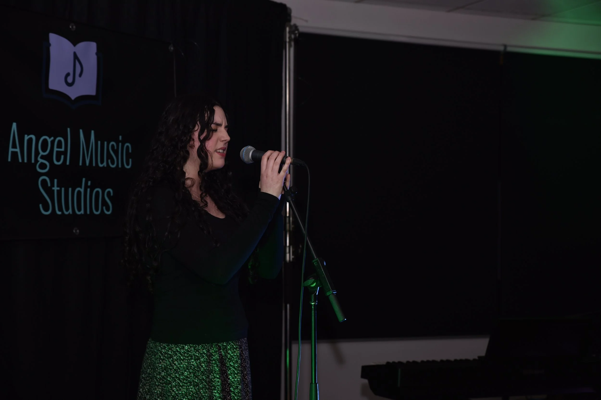 A woman with curly dark hair singing into a microphone on a stage at Angel Music Studios, with a black background and a logo of a book with a music note on it.