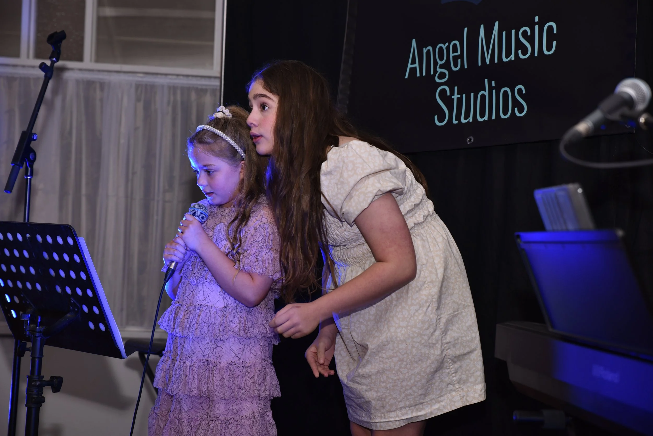 A young girl holding a microphone and a woman leaning over her, both on stage at Angel Music Studios, with music stands and microphones nearby.