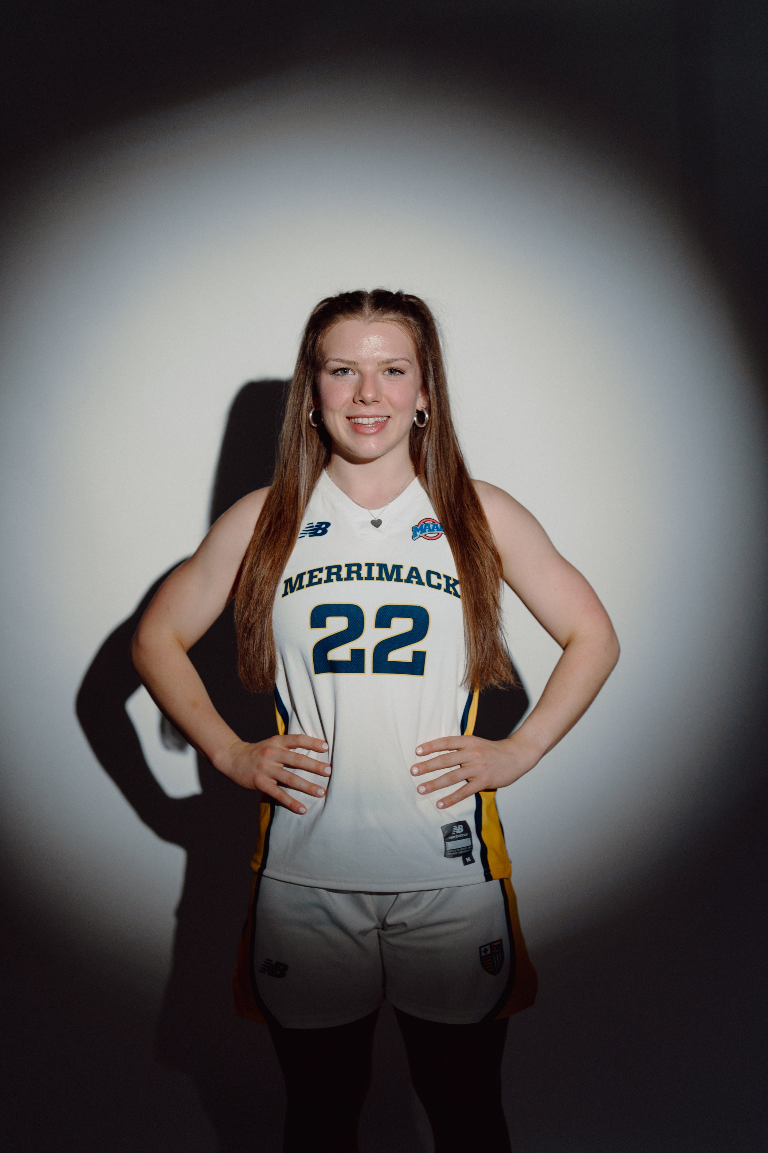 Young woman in a white sports uniform with yellow and navy accents, standing with hands on hips against a white background with a circular spotlight effect.