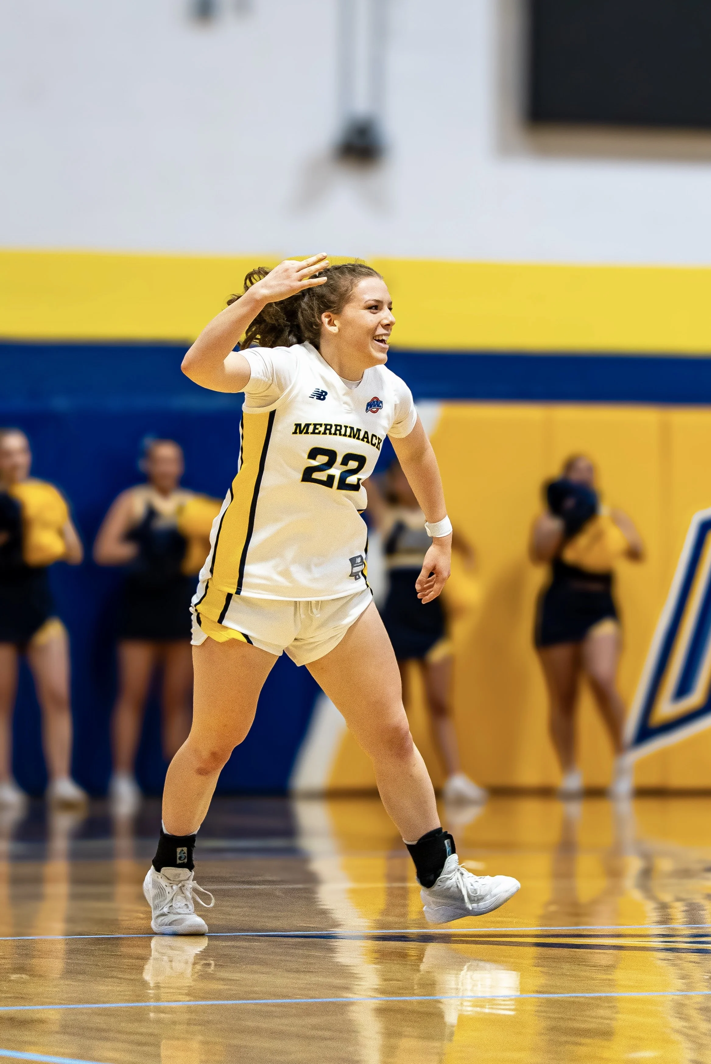 A female volleyball player with curly hair wearing a white jersey with yellow and black accents, number 22, during a game on an indoor court. She appears to be celebrating or gesturing with her right hand, with others in yellow and black uniforms blurred in the background.