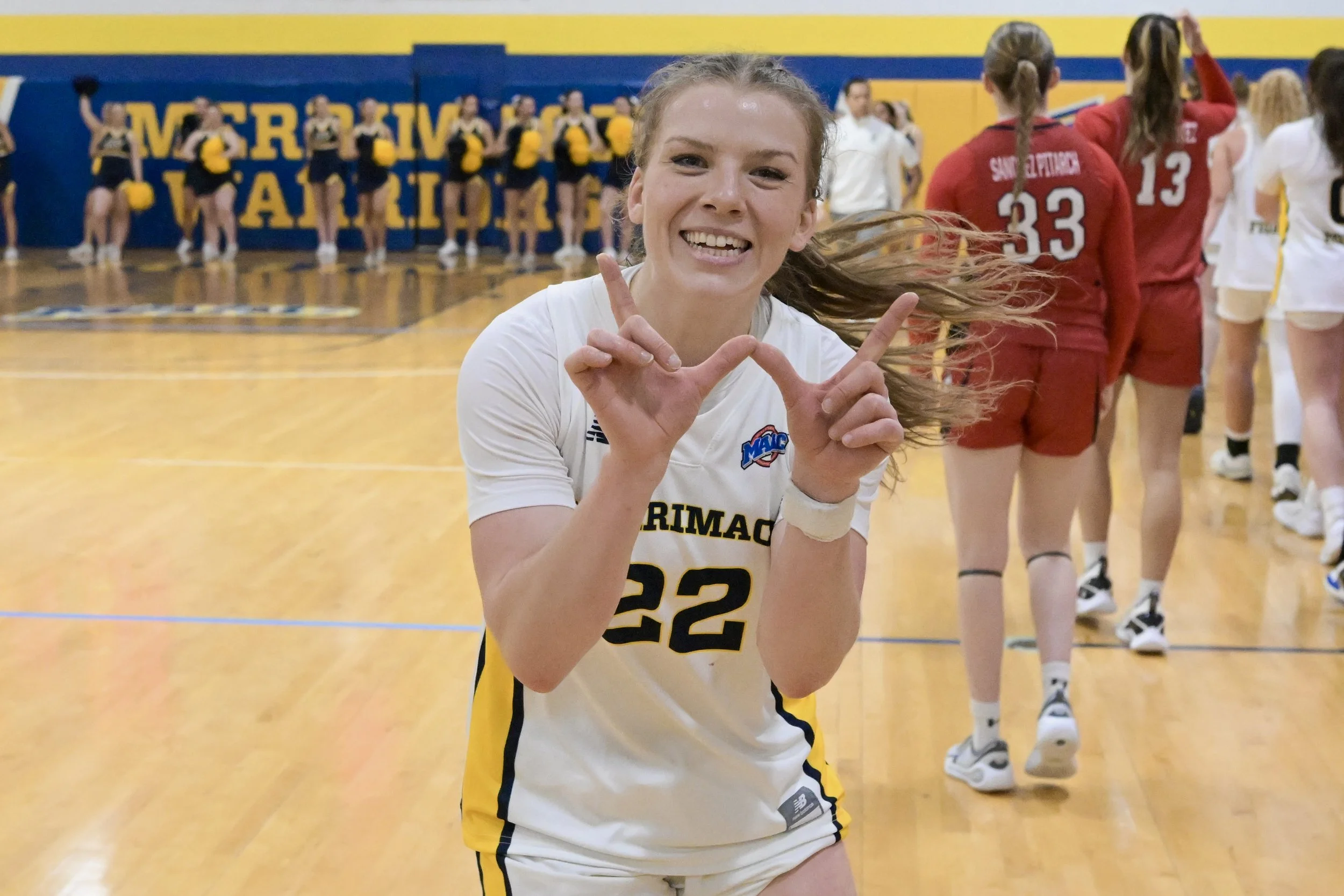 A young female volleyball player smiling and making a hand gesture on an indoor volleyball court with her teammates in the background.