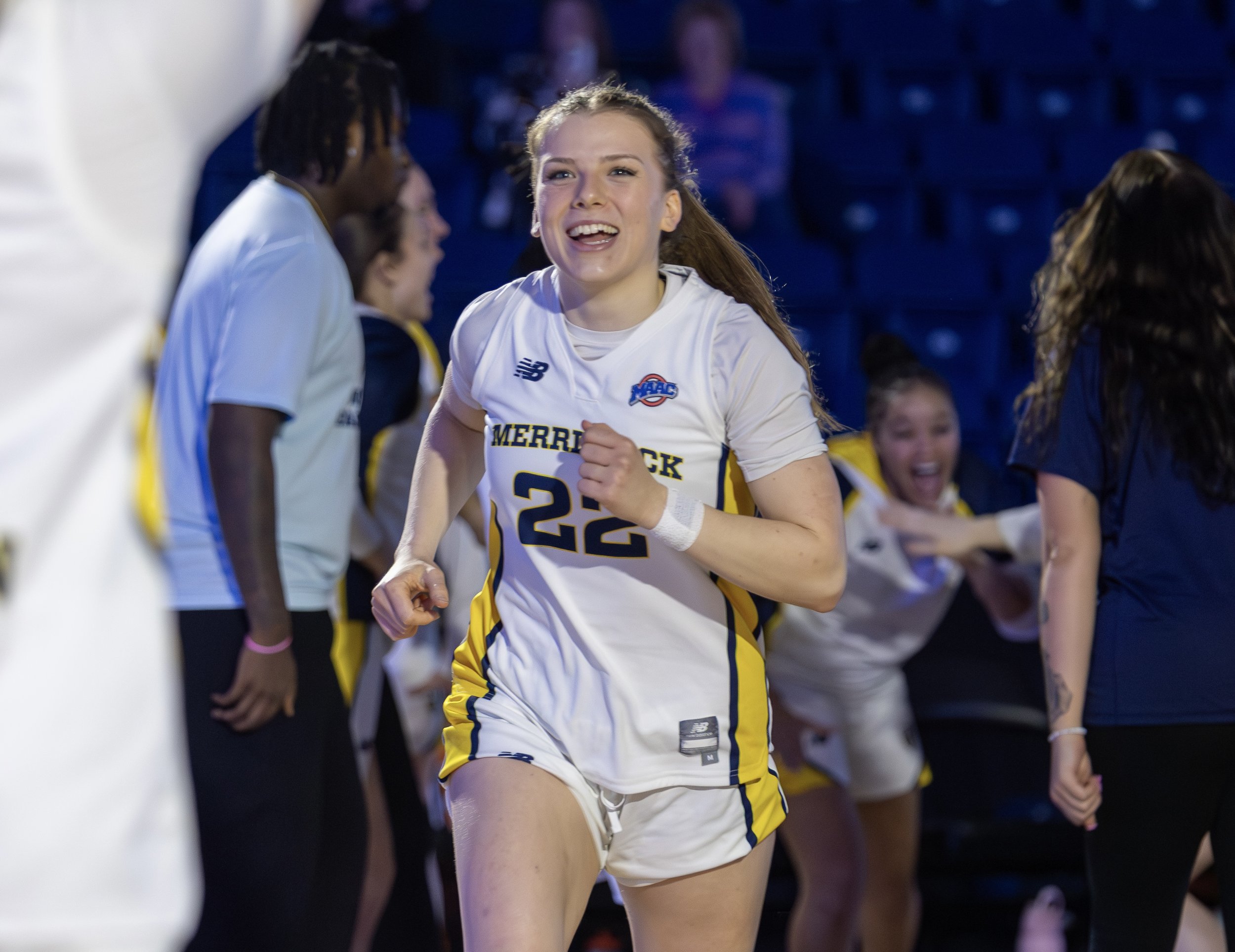 A female athlete in a white sports uniform celebrating with a smile after a game, with her teammates in the background.