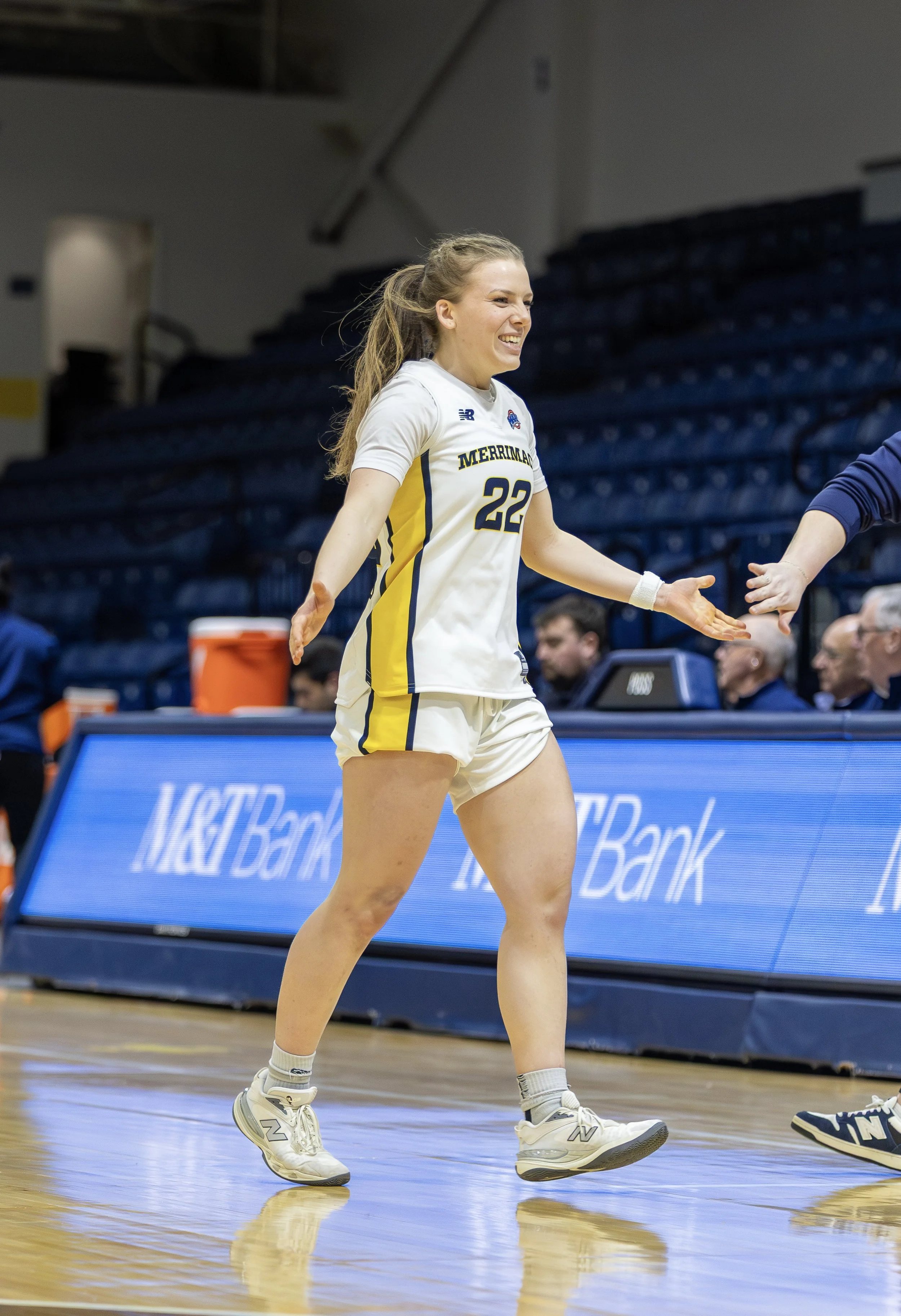 A female basketball player in a white and yellow jersey with the number 22 from Merrimack College, smiling and shaking hands during a game in a gymnasium.