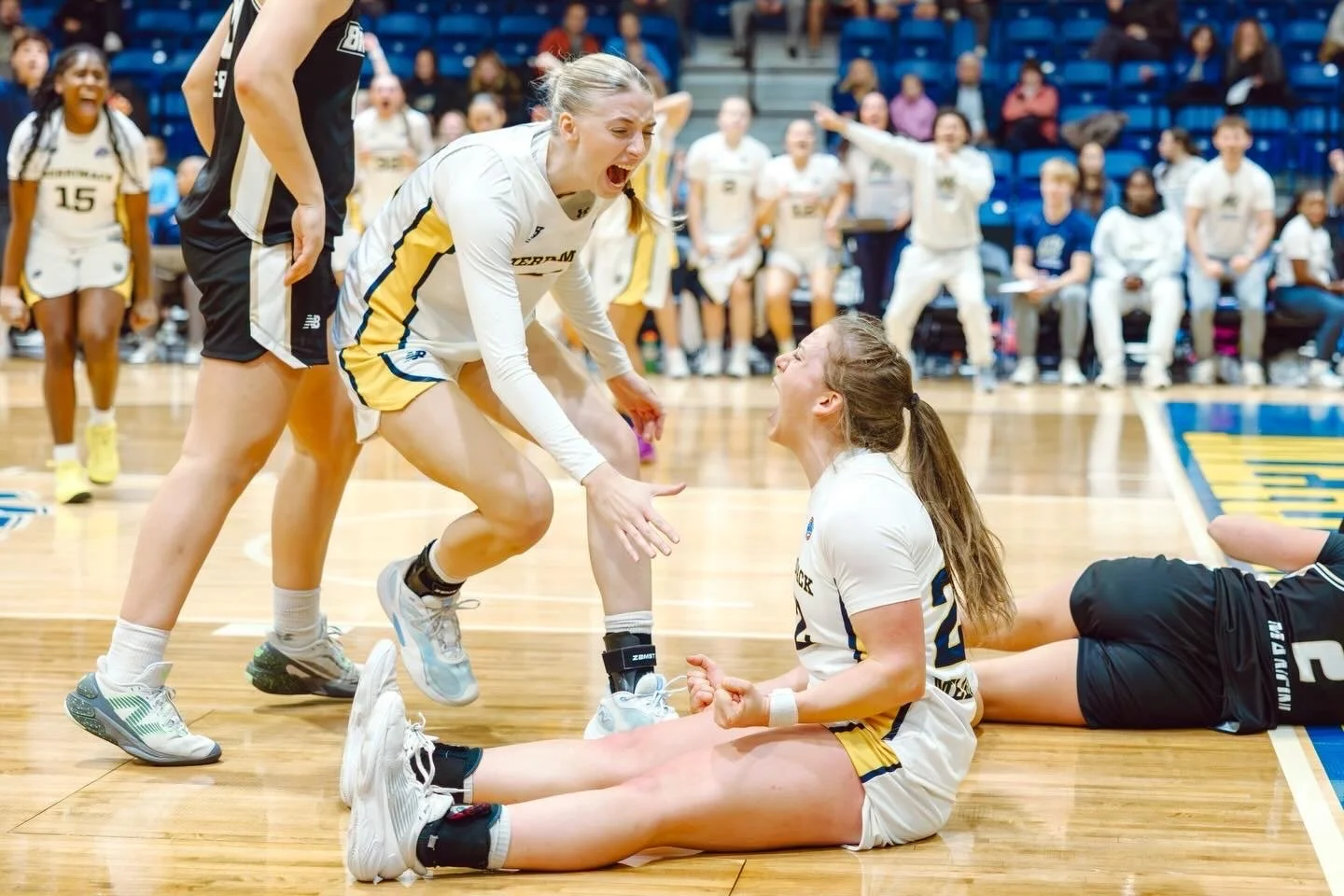Two female volleyball players are celebrating on the court; one is kneeling and smiling, while the other is leaning over, reaching out with a joyful expression. A third player is lying on the floor nearby, and cheering players and spectators are visible in the background.