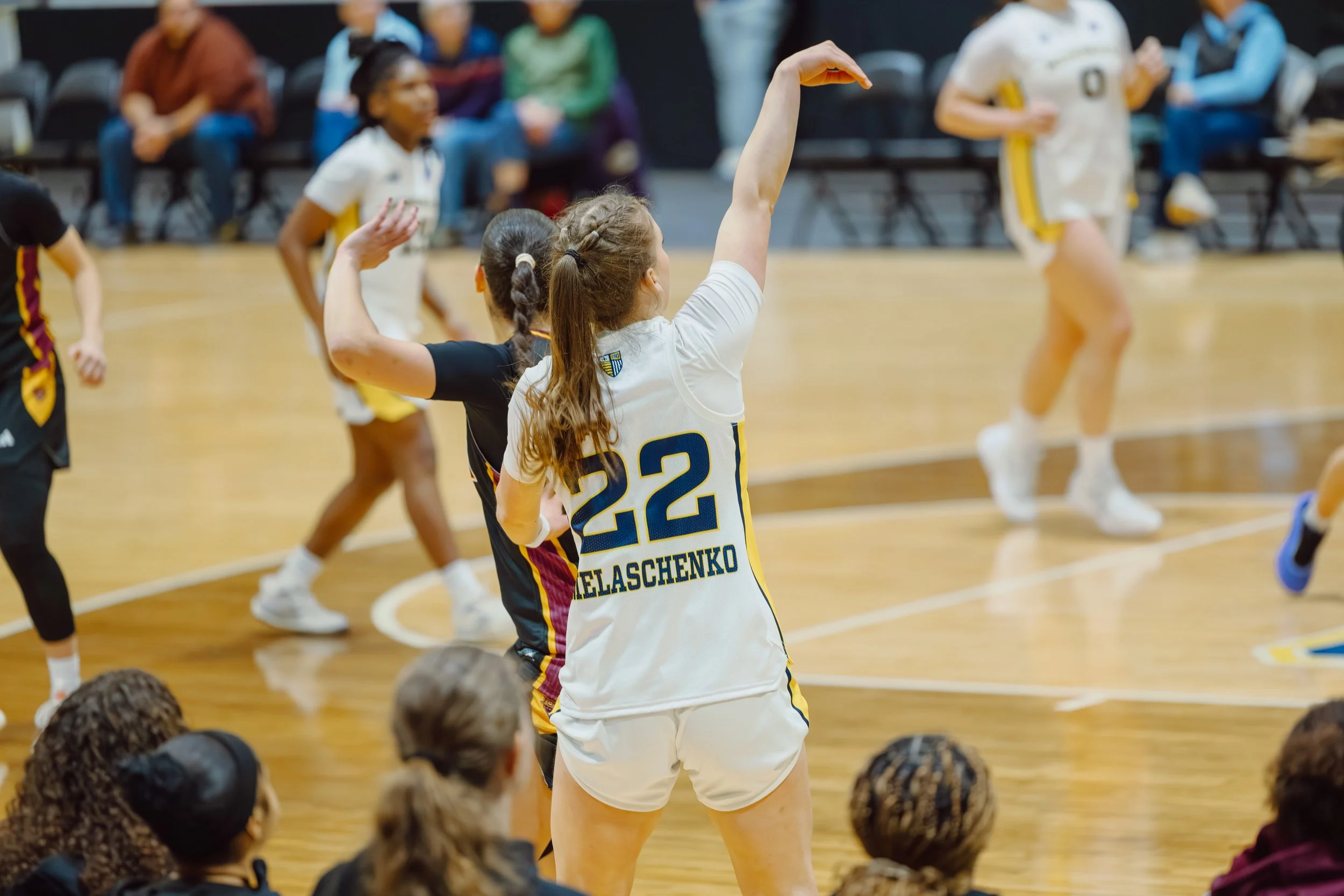 A female basketball player with jersey number 22 practicing free throws on a basketball court, while spectators and other players observe in the background.