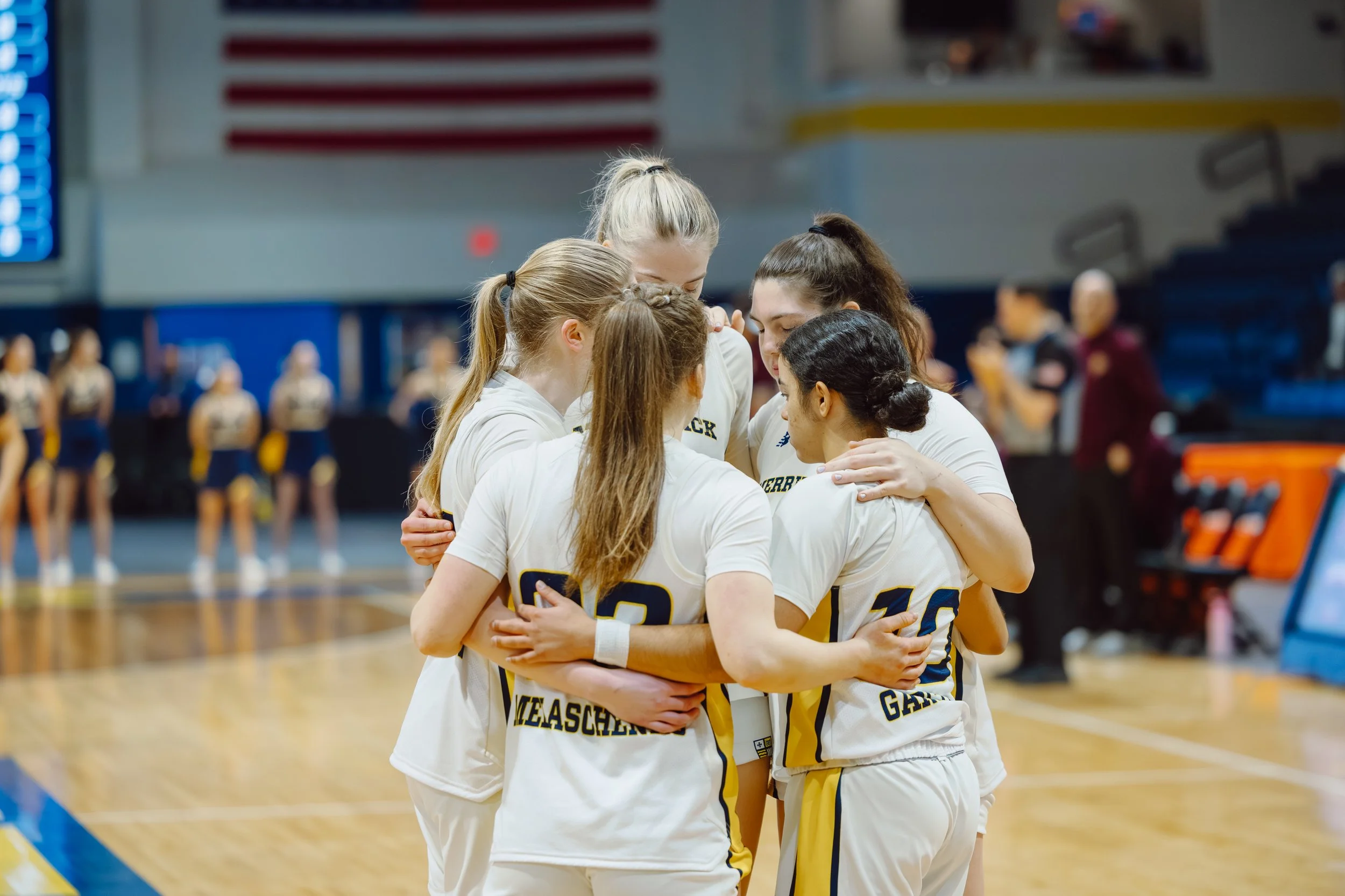 A group of young female volleyball players in team uniforms huddle together in a gymnasium during a game or practice, with a volleyball court, court lines, and other players visible in the background.