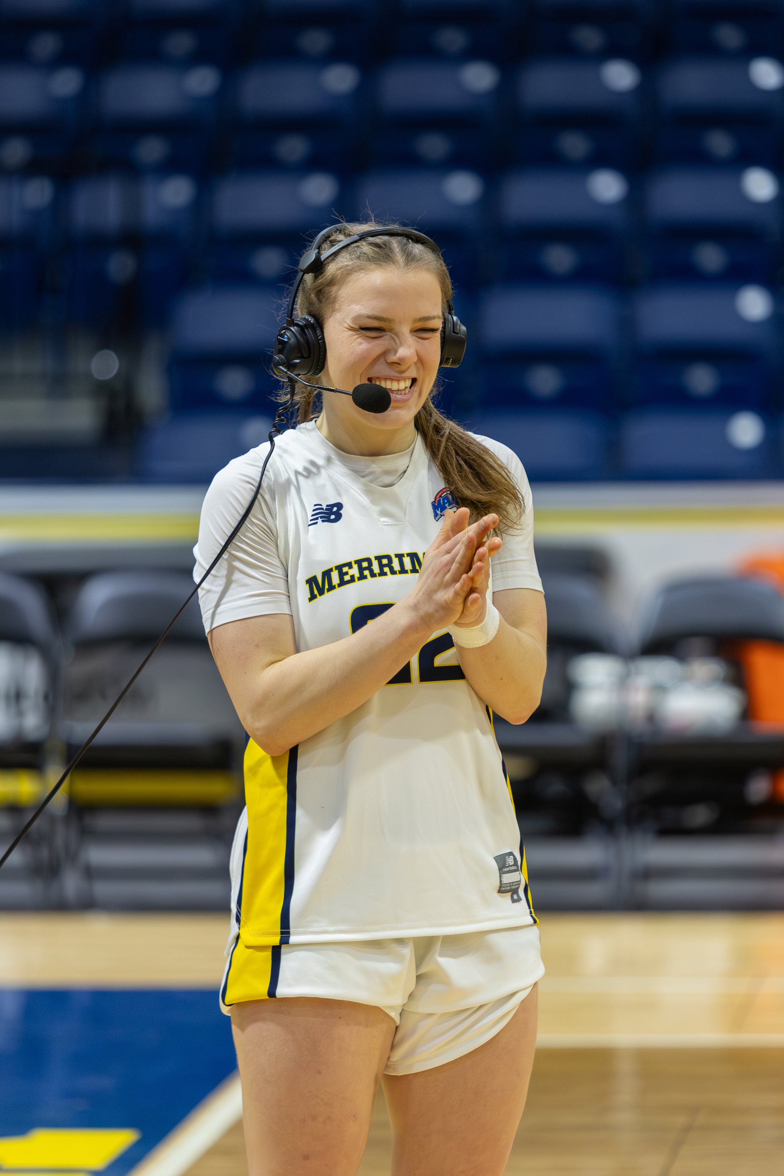 A female volleyball player in a white and yellow uniform with the text 'MERRIMAC' on her jersey, smiling and clapping her hands, wearing a headset, on an indoor court.