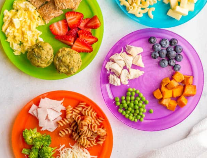 Colorful plates with various foods including strawberries, blueberries, shredded chicken, carrots, green peas, pasta, and cheese, arranged on a white surface.