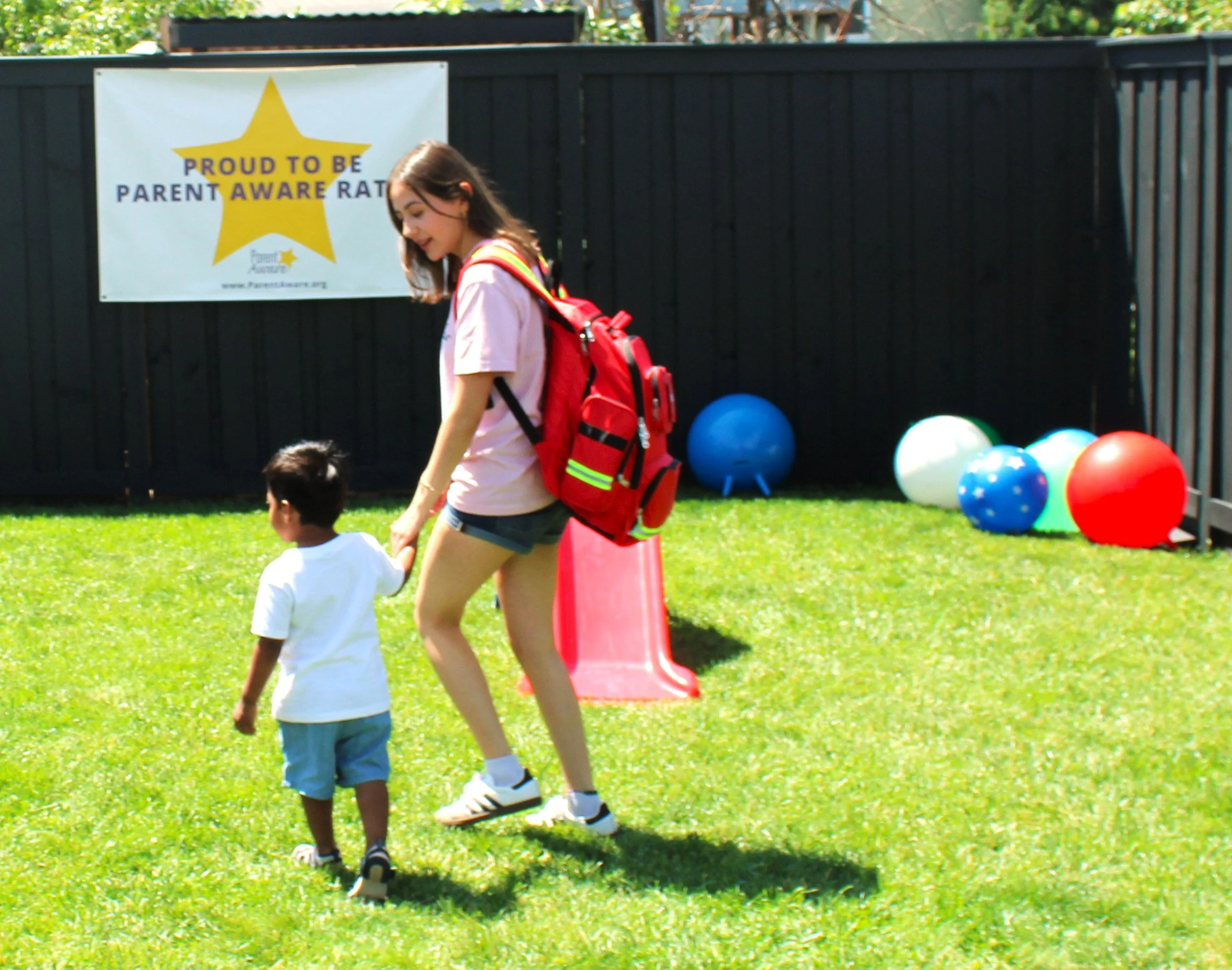 A young girl wearing a pink shirt, shorts, and sneakers is outdoors on a grassy area. She has a red backpack and is walking past a boy who is also outside. There are colorful balloons on the ground and a black fence in the background with a sign that reads 'Proud to be Parent Aware Rated'.