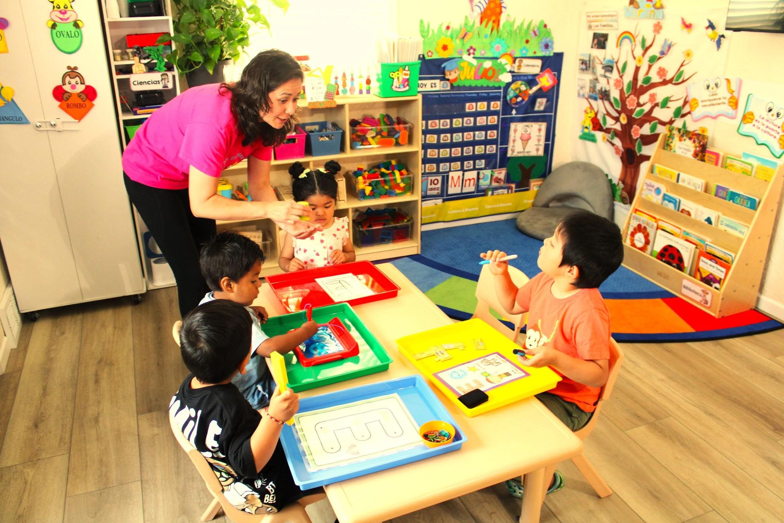 A teacher with dark, curly hair wearing a pink shirt teaches a group of young children seated at a table in a colorful classroom. The children are engaged in activities with trays of supplies, and the classroom features vibrant educational decorations, bookshelves, and artwork on the walls.