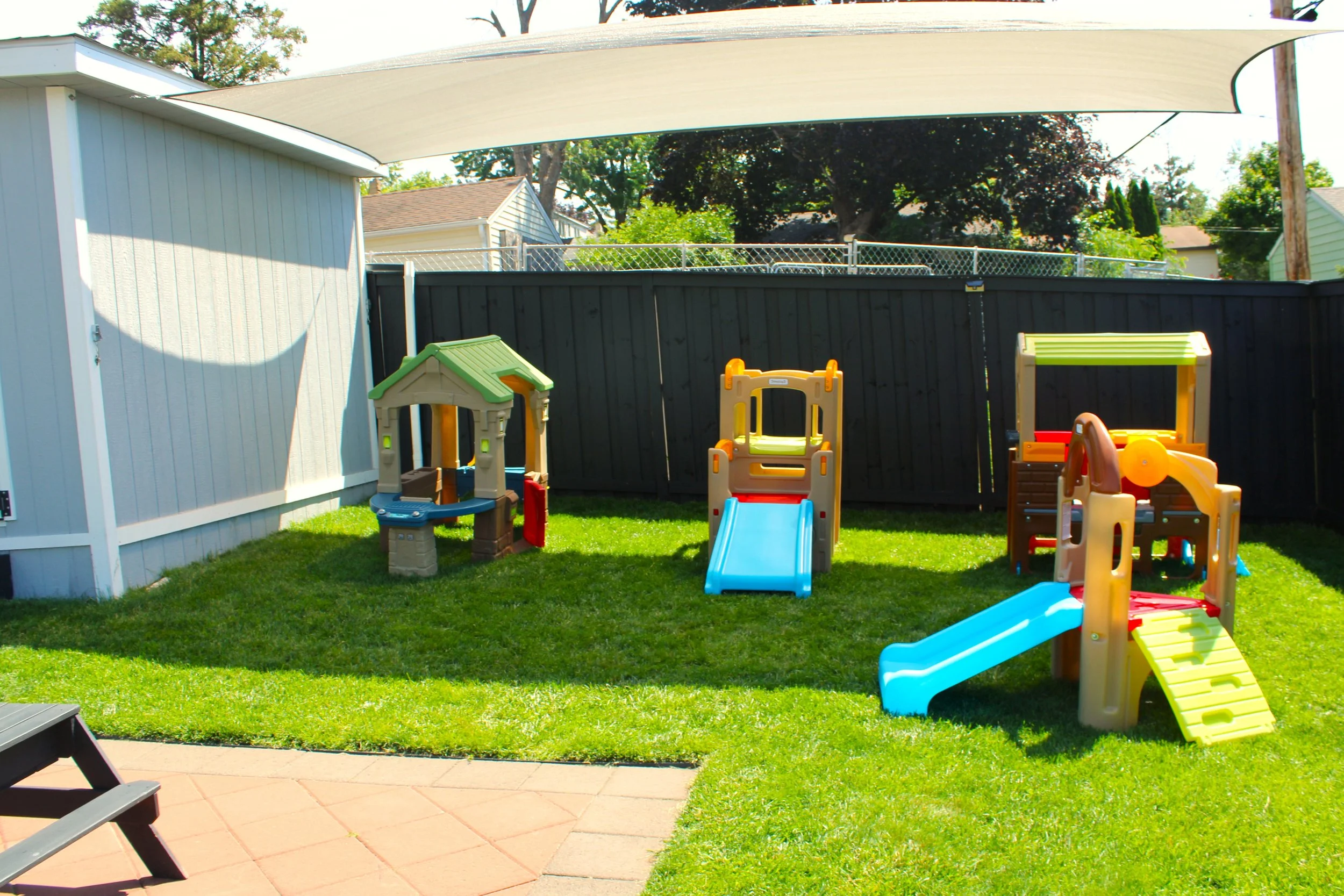 Child's backyard playground area, with small slides, a playhouse, and a sandbox on a grass lawn, enclosed by black fencing, and shaded by a large shade sail.