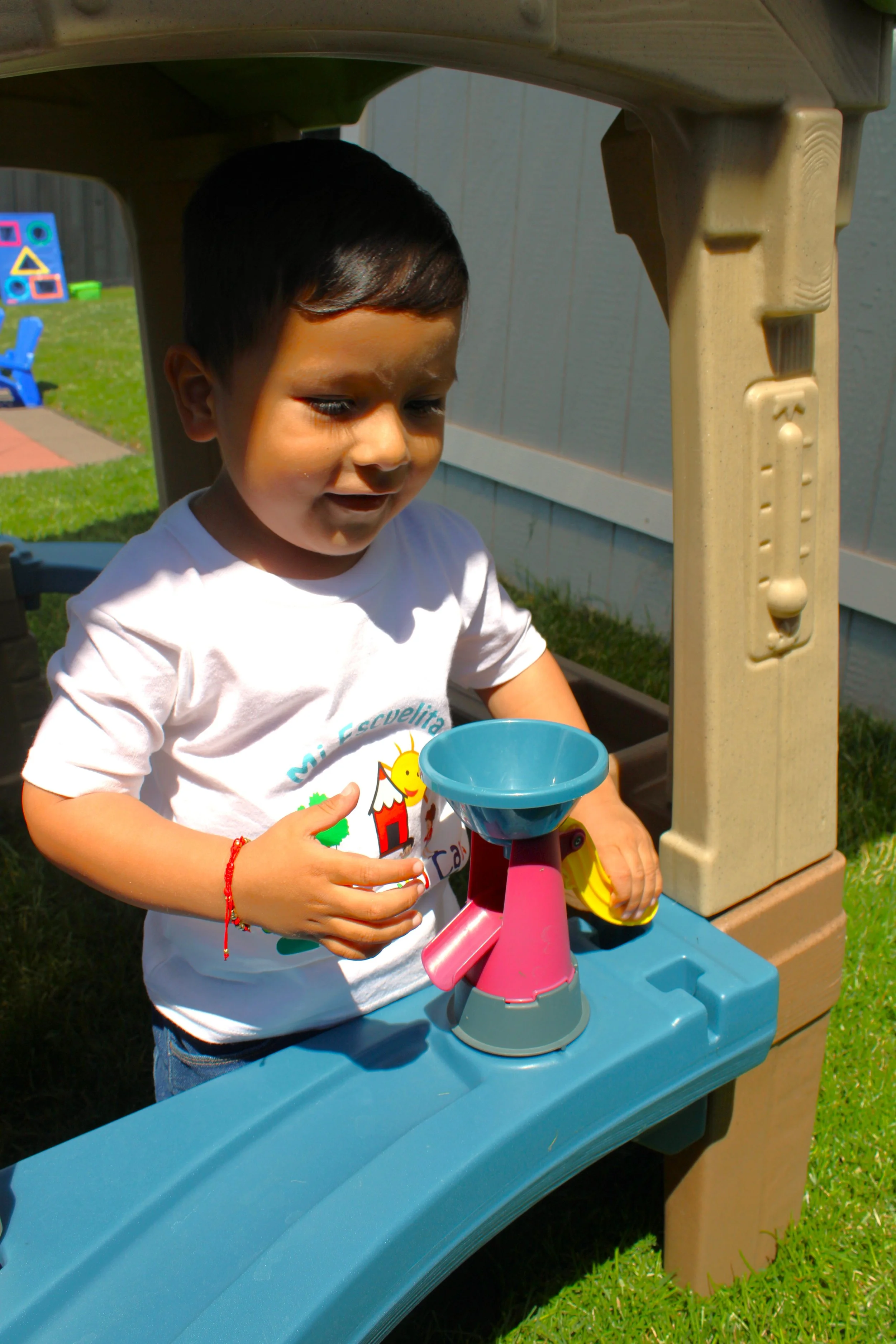 A young boy with dark hair and a white t-shirt playing with a toy outdoor kitchen set, with a slide and a grassy yard in the background.