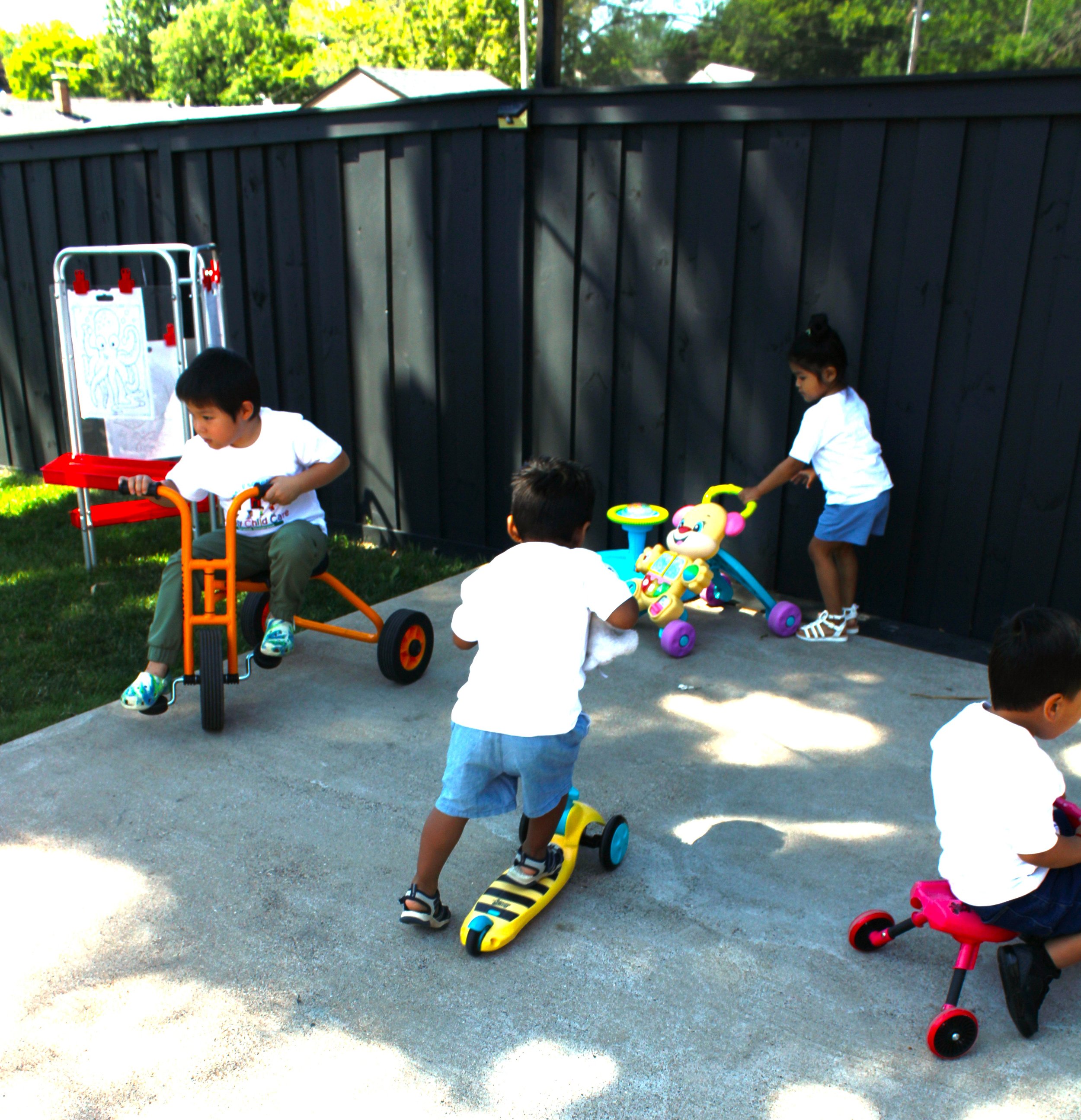Children playing outside with scooters and toy push cars in a backyard enclosed by a black fence, with trees and houses in the background.