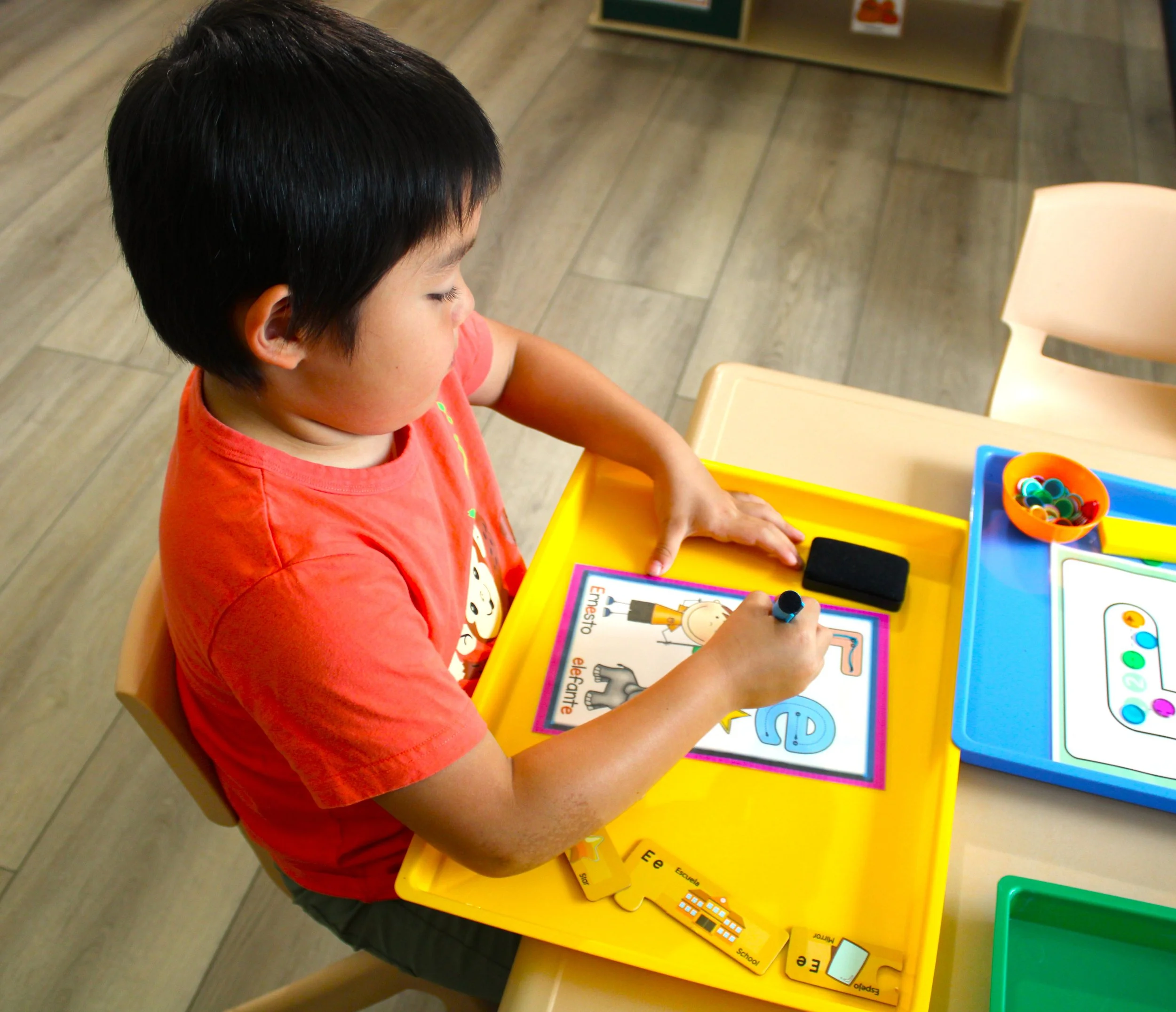 A young boy with dark hair wearing a red shirt sitting at a yellow table playing with educational toys and illustrated learning materials.
