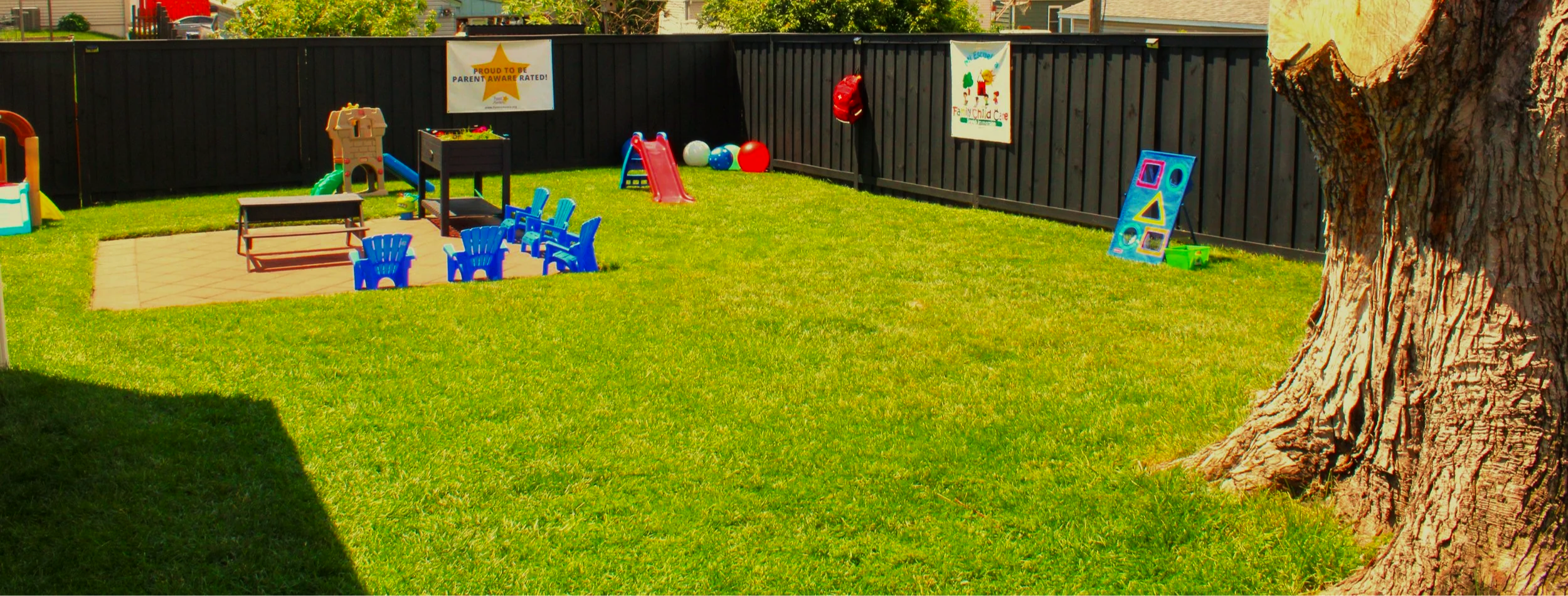Children's outdoor playground area with toys, chairs, and a sandbox, surrounded by a black fence and a large tree.