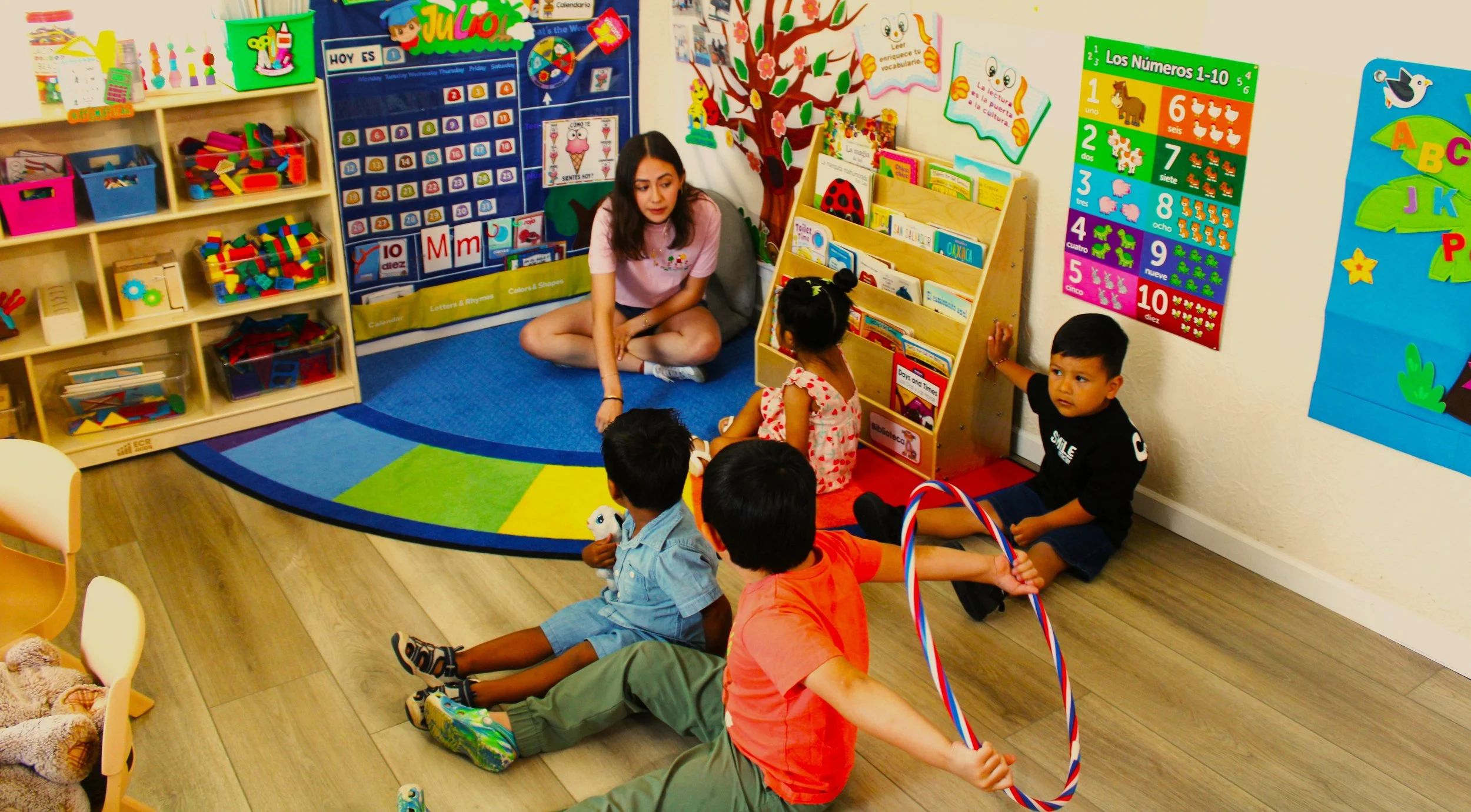 A group of young children and a woman are sitting on the floor in a colorful classroom, engaged in a group activity or storytime. The room is decorated with educational posters, bookshelves, and bright, educational decorations.