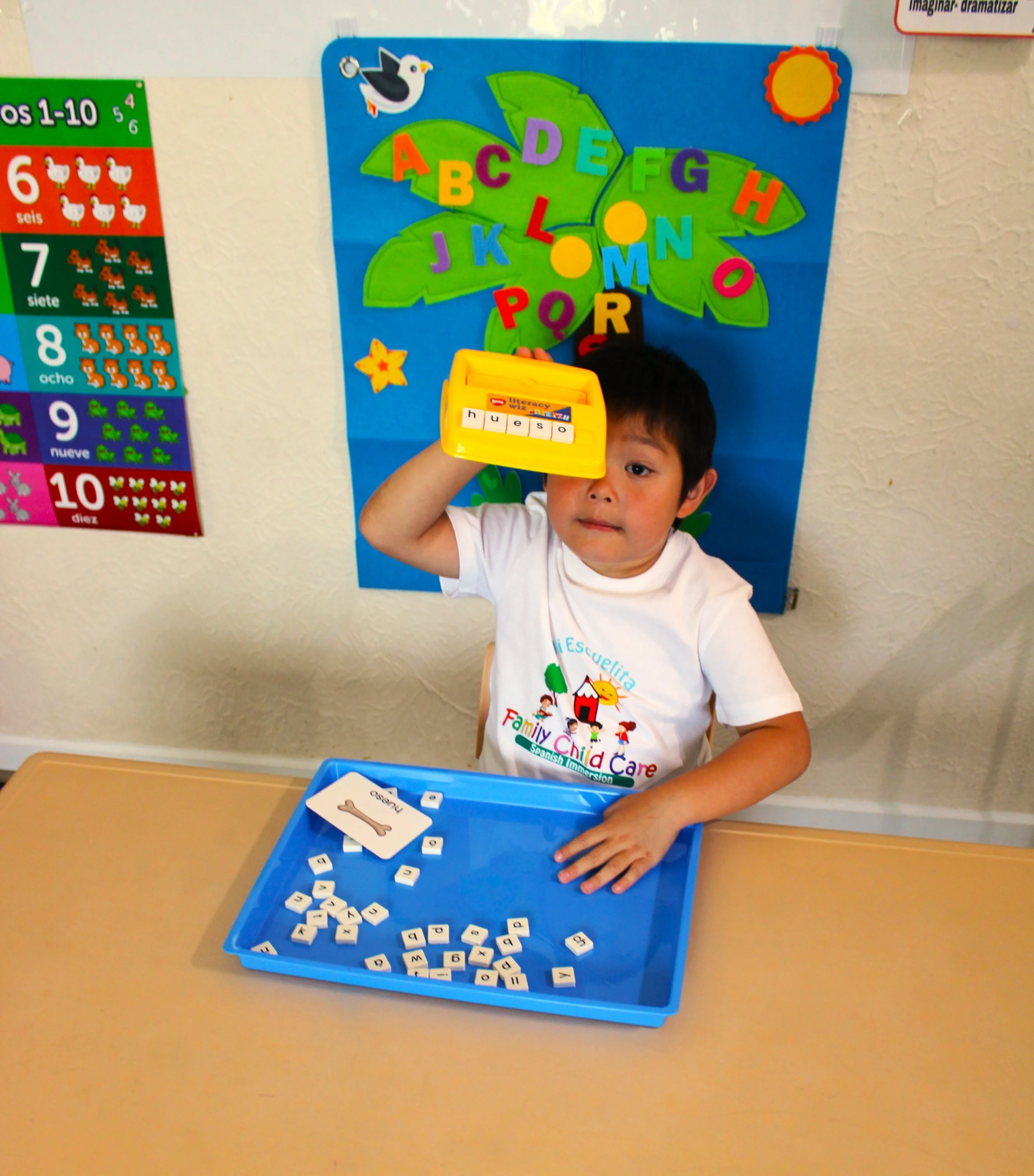 A young boy holding a yellow box of letter tiles and playing with white letter tiles in a blue tray on a table, in a classroom with educational posters on the wall.