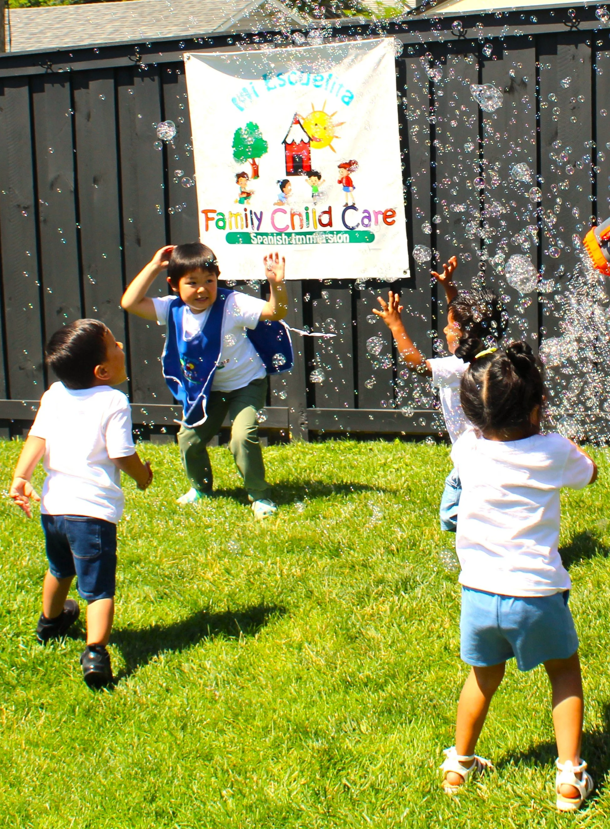 Children playing with soap bubbles outdoors in front of a black fence with a colorful banner that reads 'Valle Escondida Family Child Care'.