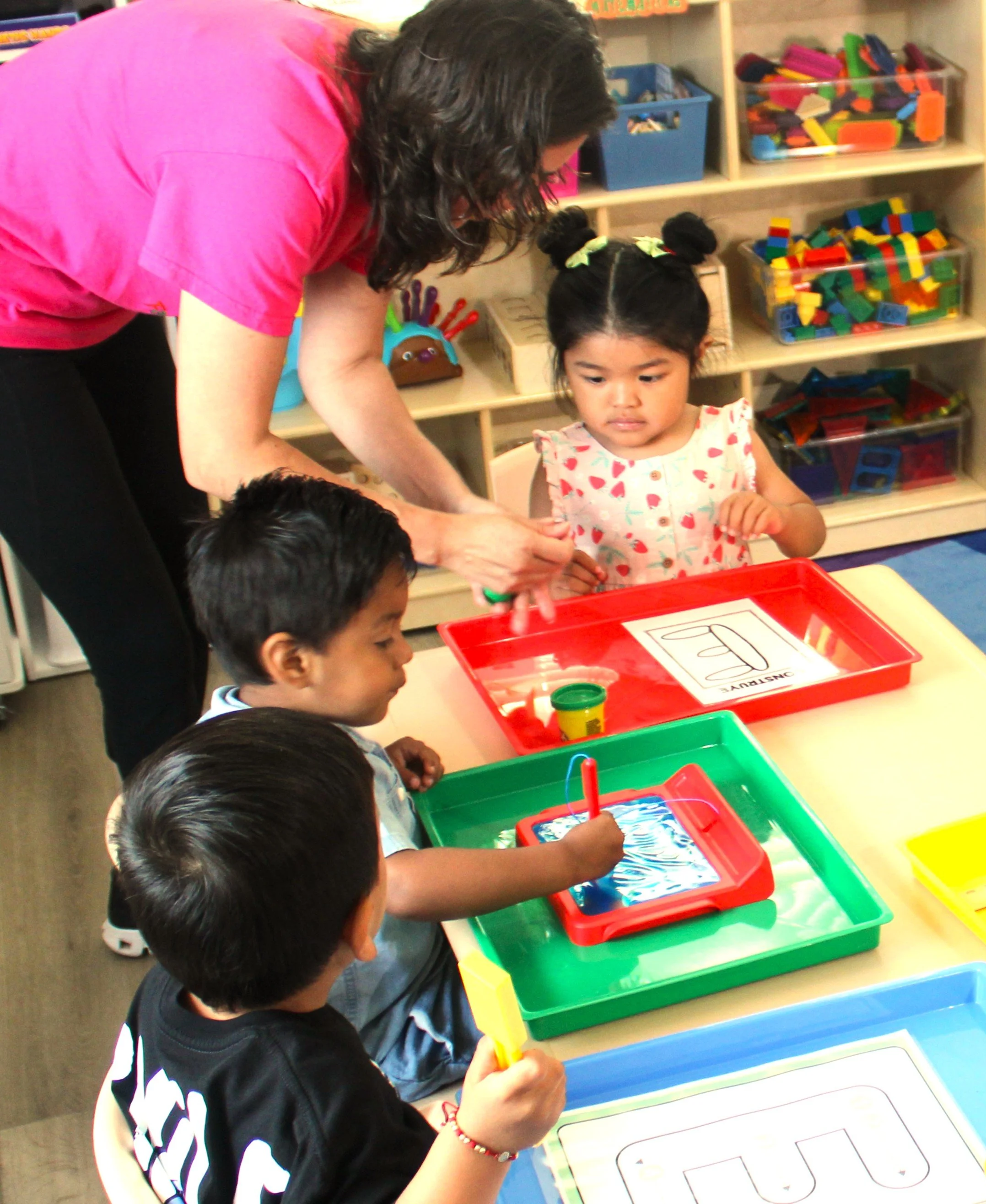 Children engaging in a hands-on learning activity at a table with colorful bins filled with building blocks and drawing sheets in a classroom setting.
