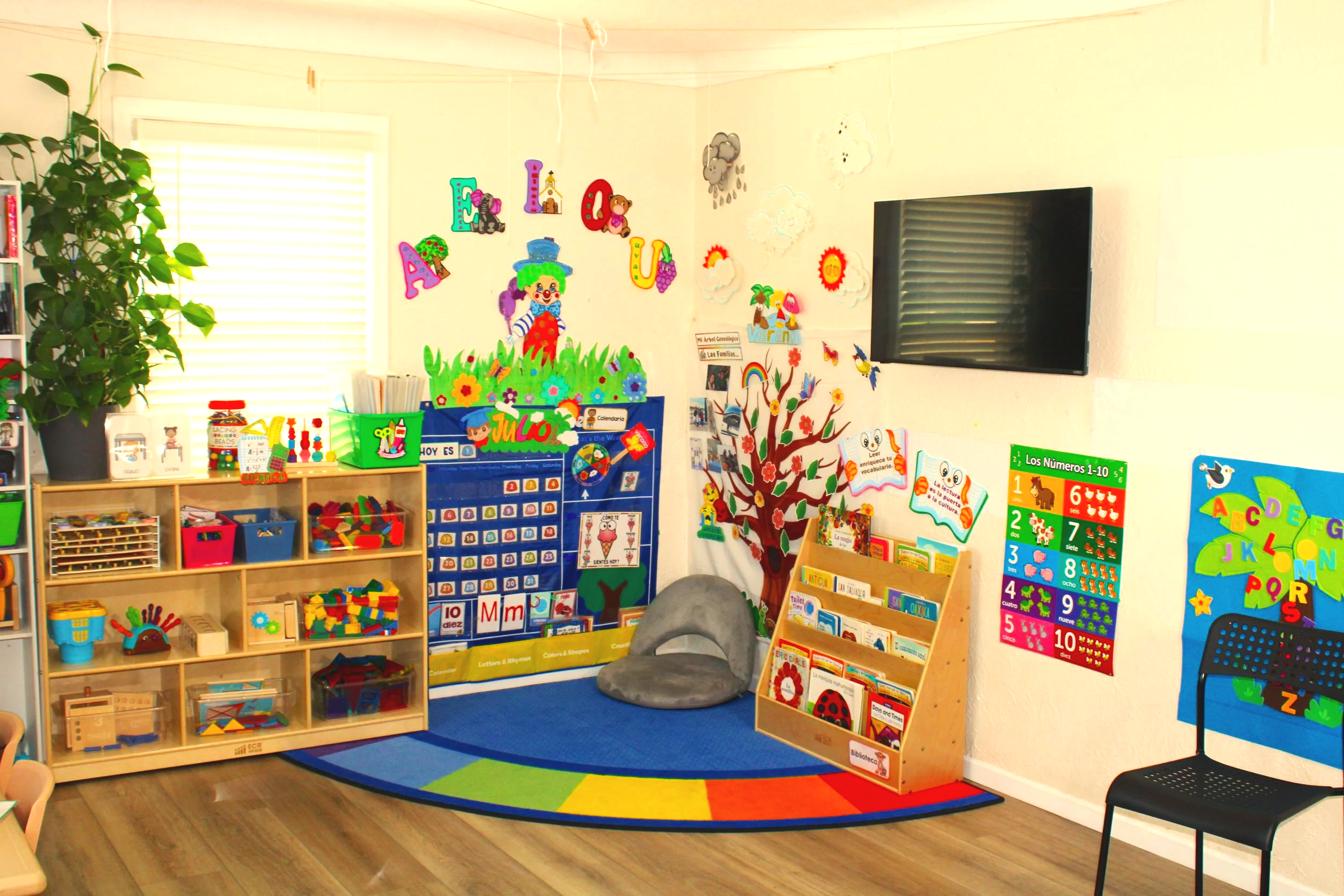 Colorful classroom corner with educational posters, bookshelves filled with educational toys and books, a vibrant rainbow carpet, and decorative wall art including clouds, animals, and the alphabet.