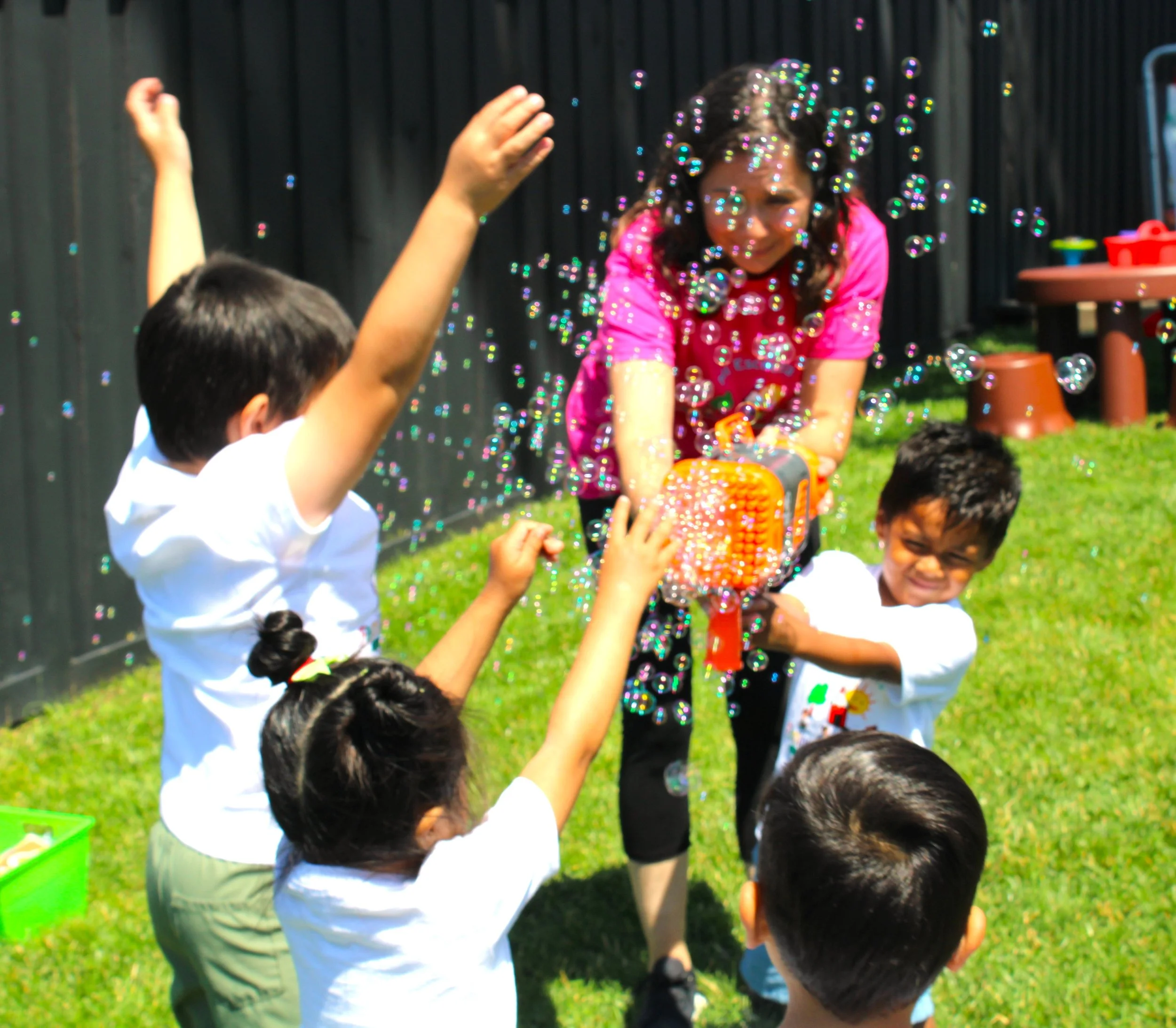 Children playing outdoors with bubbles on a sunny day, with a girl in a pink shirt and several other children reaching towards the bubbles.