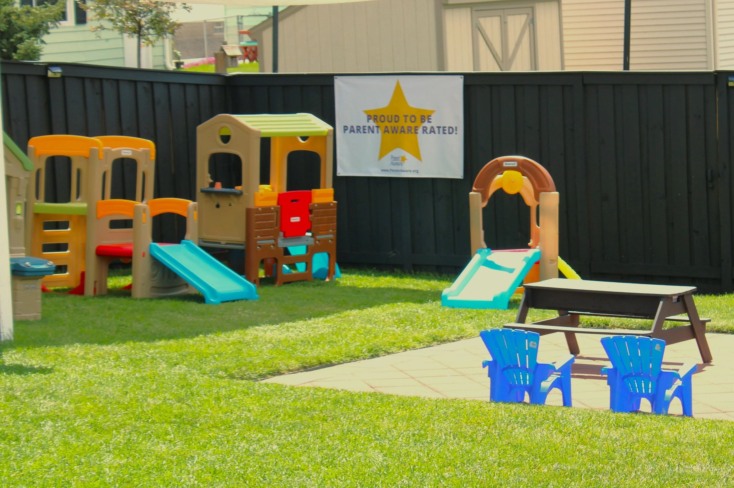 Children's playground with slides, a small house, and blue chairs, enclosed by a black fence. A banner on the fence reads 'Proud to be parent aware rated!'