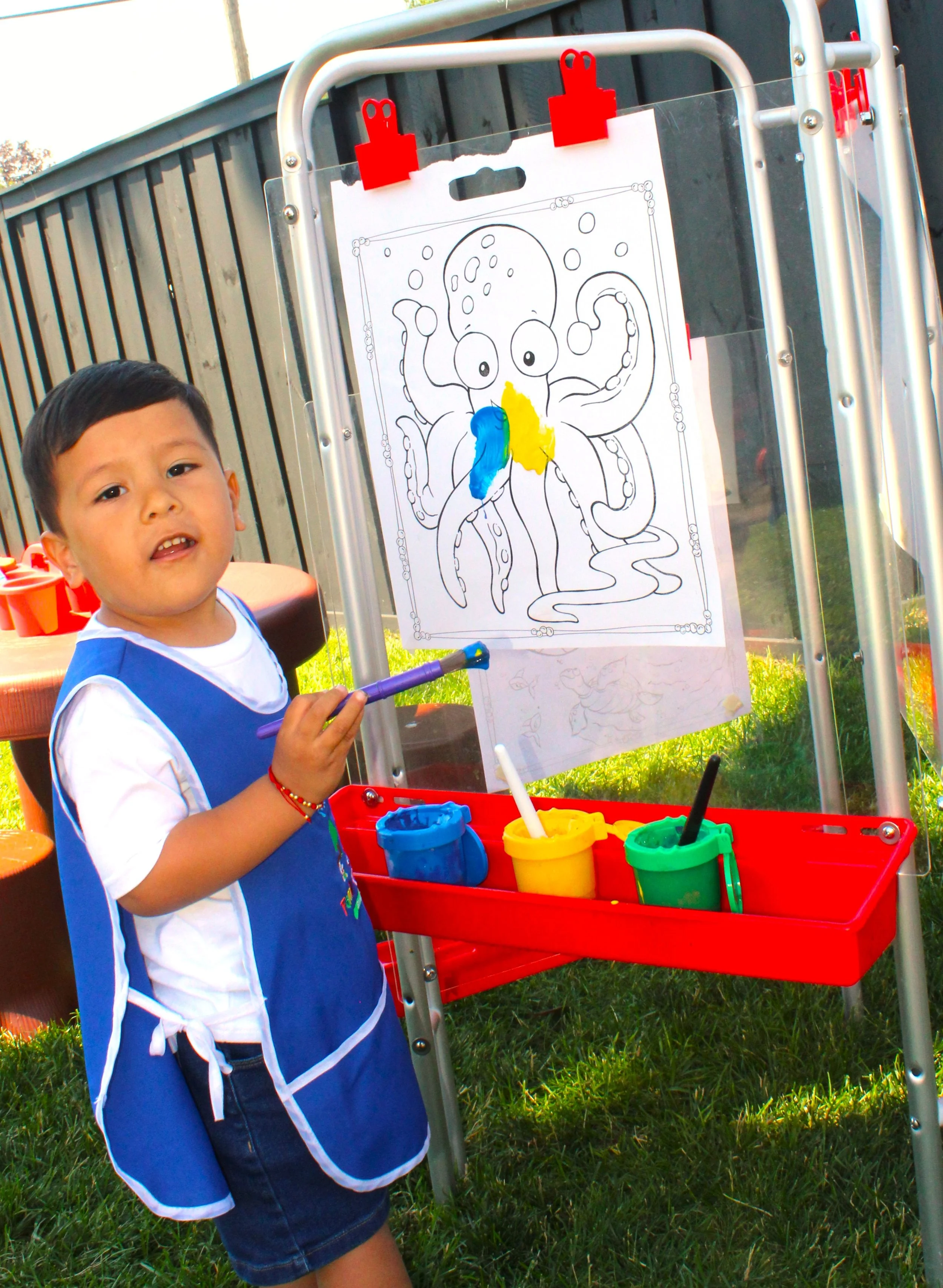 A young boy with short black hair wearing a blue apron over a white t-shirt and denim shorts, standing outside on grass, holding a purple paintbrush, next to an easel with a coloring page of an octopus. The octopus coloring page is partially colored with blue and yellow sections. There are containers of yellow, green, and blue paint on a red tray attached to the easel.