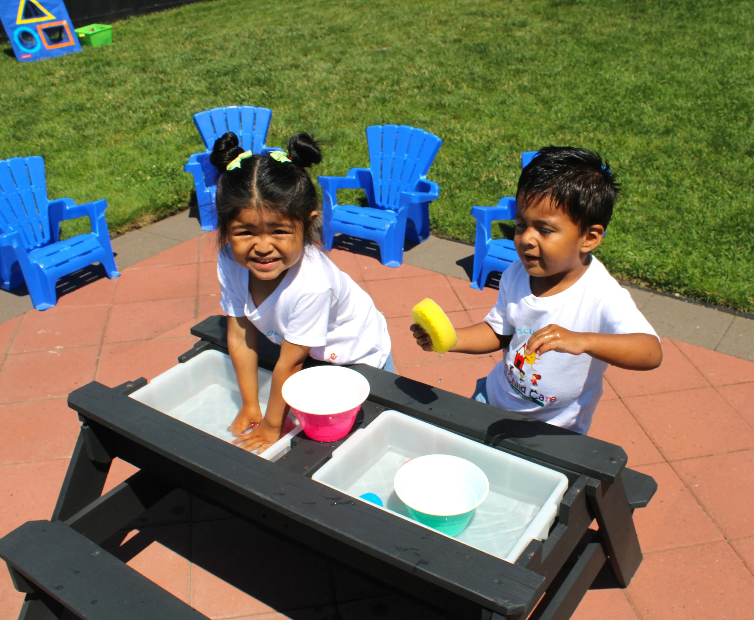 Two children are playing at a water table outdoors on a sunny day, surrounded by green grass and blue Adirondack chairs. One girl with pigtails is smiling, and a boy is holding a yellow sponge.