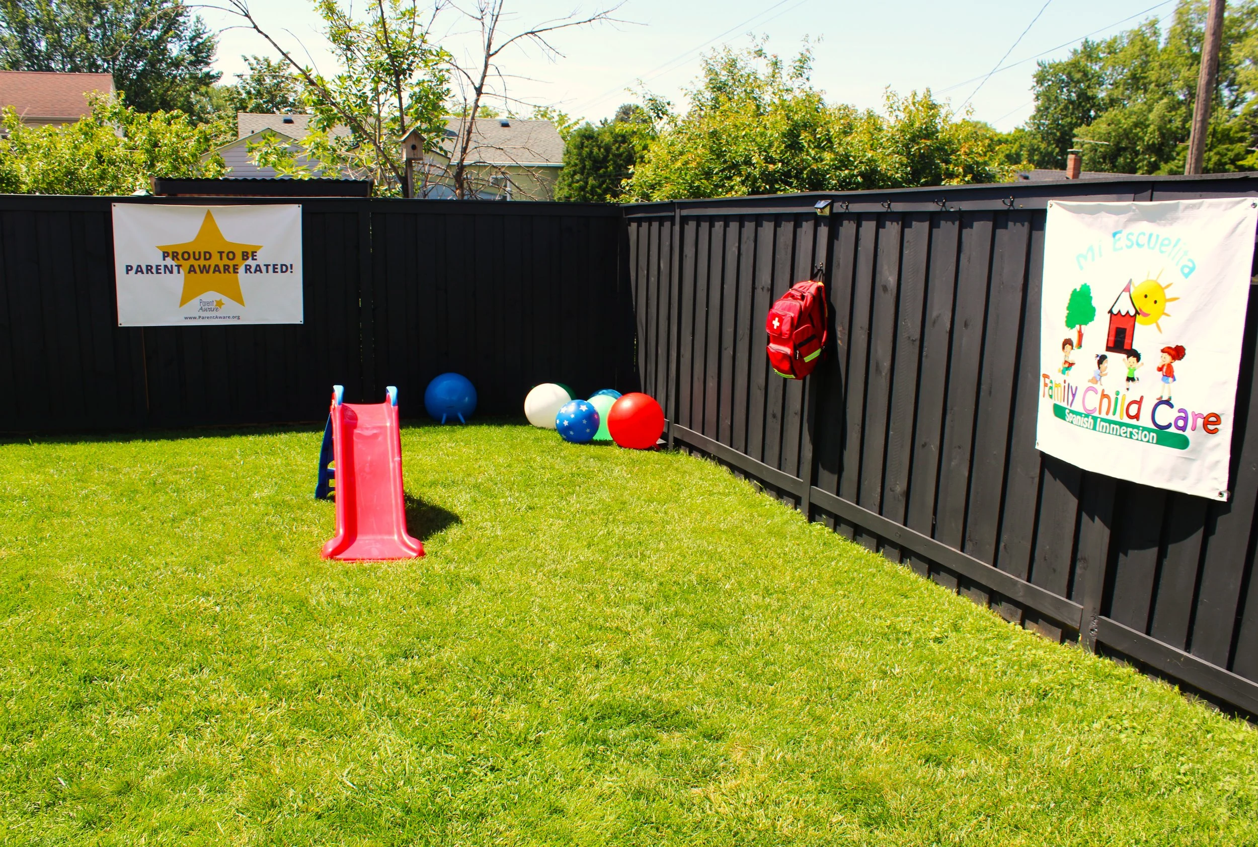 Backyard with a black fence, playground slide, colorful balls, a red backpack, and banners promoting parenting awareness and children's Spanish immersion.