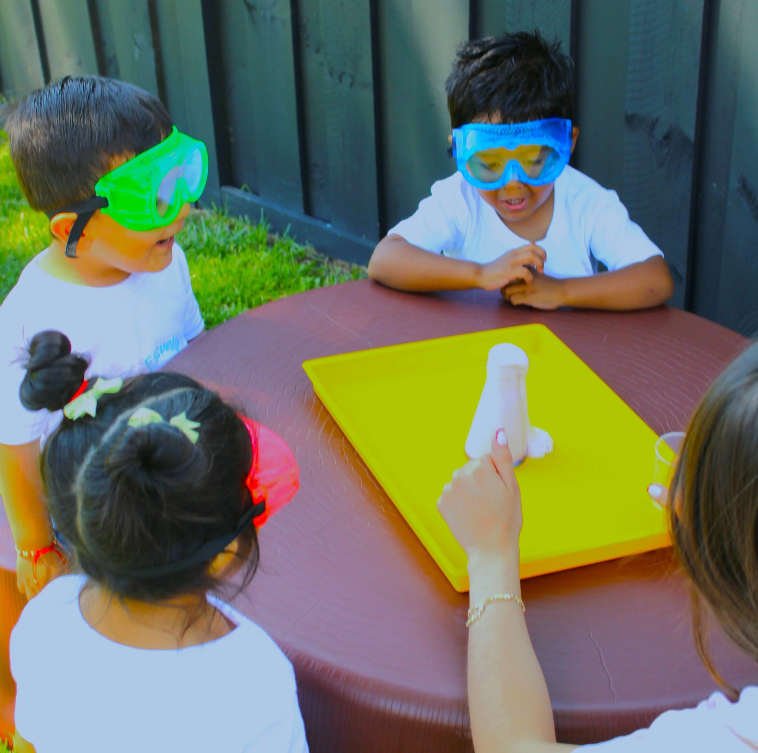 Two young boys wearing swimming goggles sitting at an outdoor table, engaging in an activity.