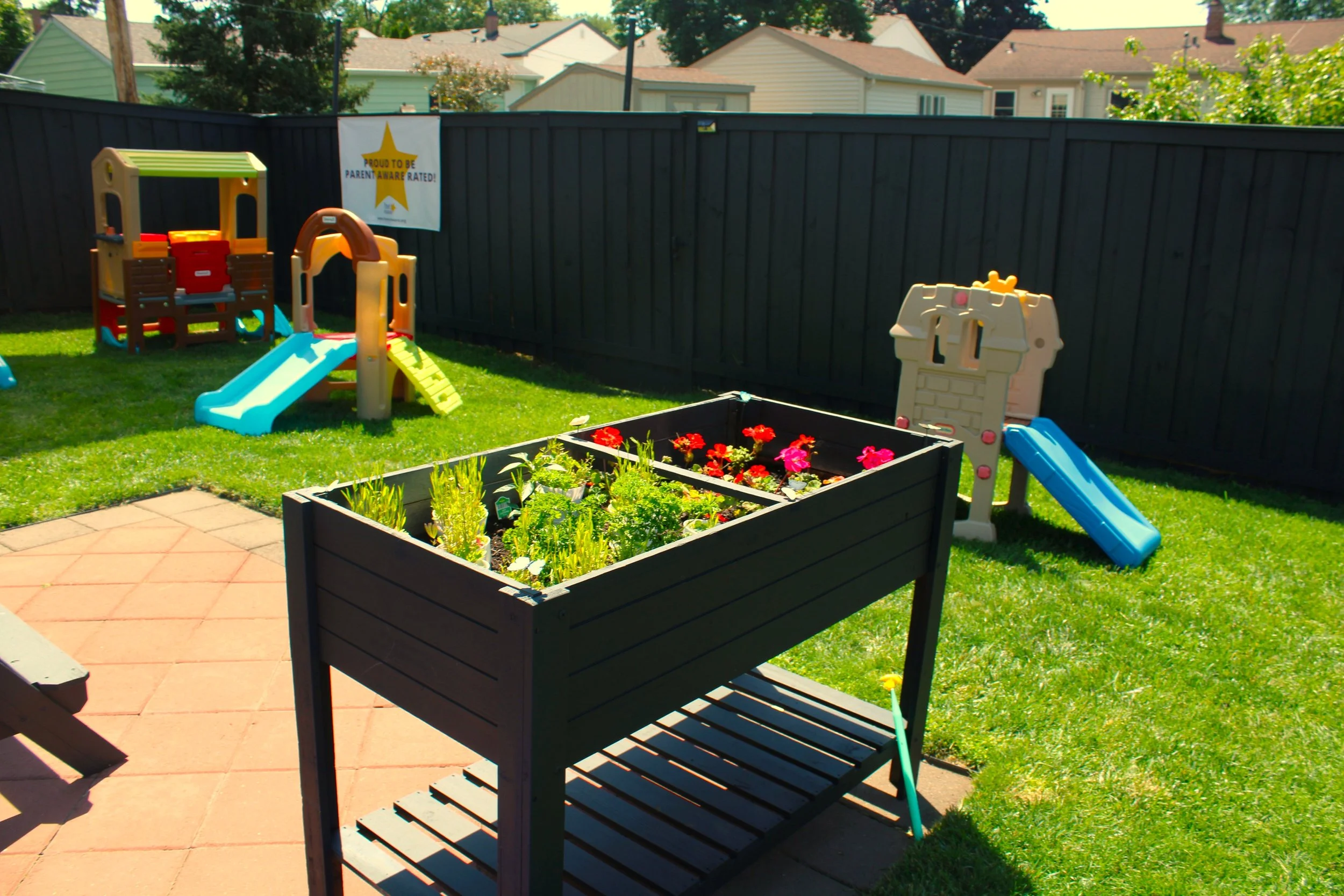 Children's backyard with plastic playhouse, slide, garden bed with flowers, and patio area, surrounded by a black wooden fence and neighboring houses.