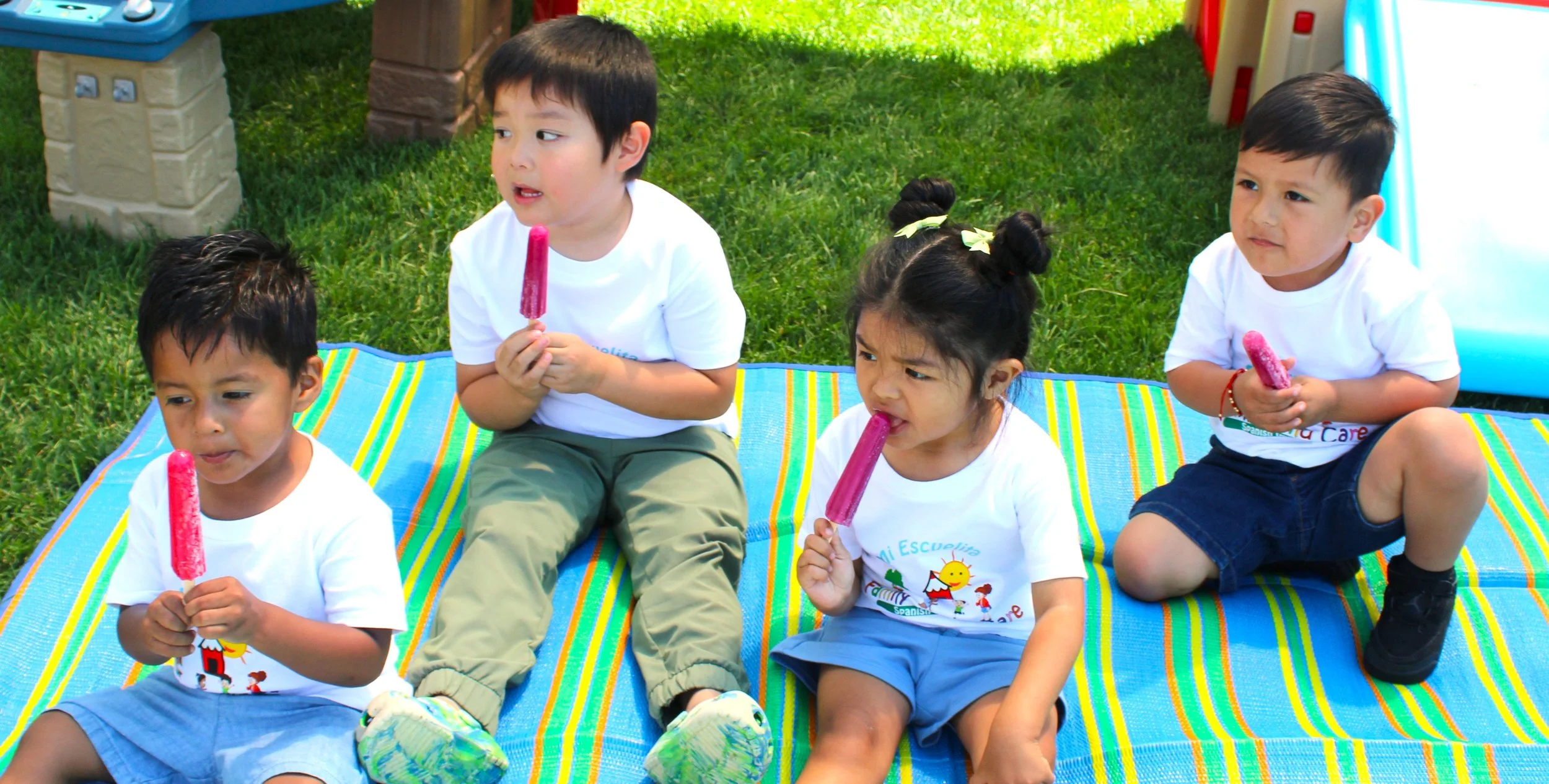Four young children seated on a multicolored striped mat outdoors, each holding a pink popsicle, in a grassy area with play structures visible in the background.