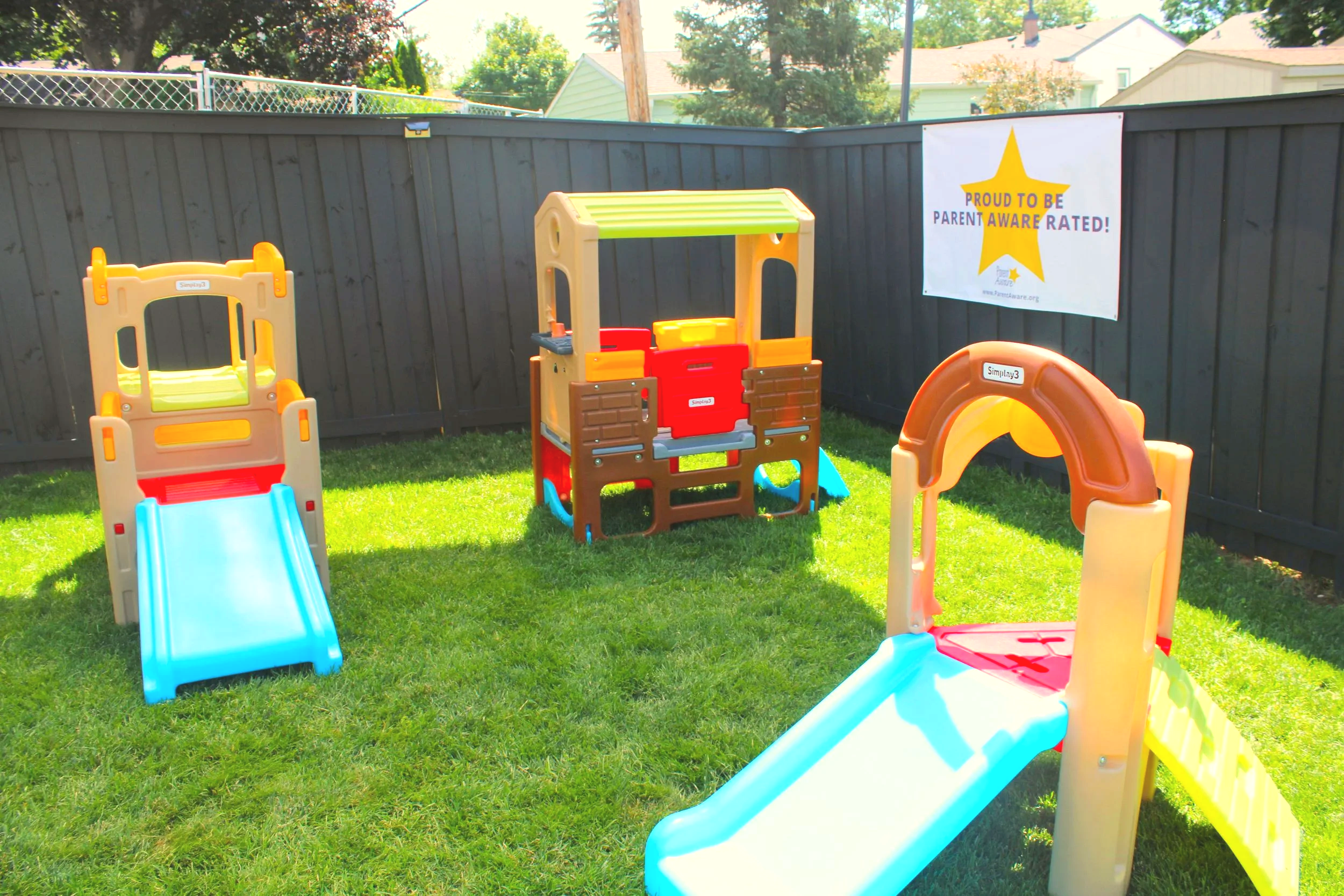 Colorful plastic playset with slides and climbing structures in a backyard surrounded by a dark wooden fence, with a banner that reads 'Proud to be parent aware rated!'