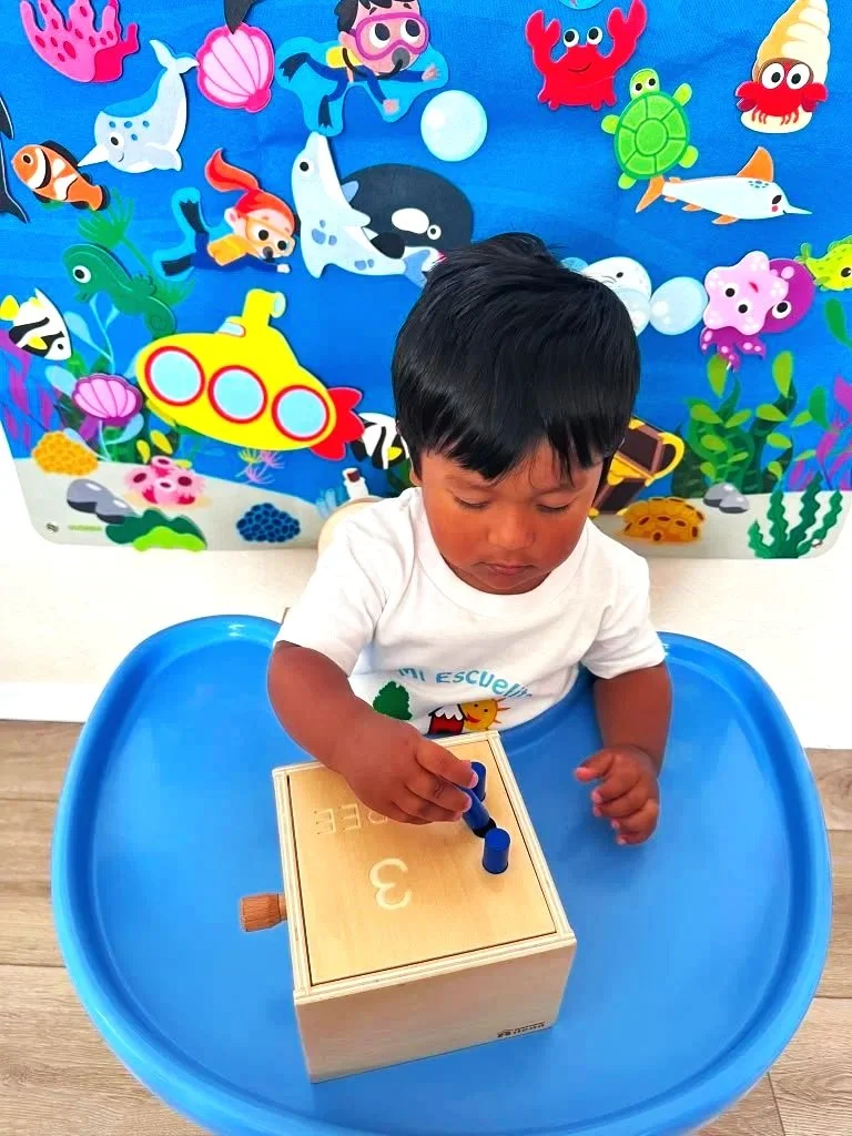A young boy sitting at a blue tray table, playing with a wooden box that has the number three and the letter 'E' on it, in front of a colorful underwater-themed backdrop with cartoon sea creatures.