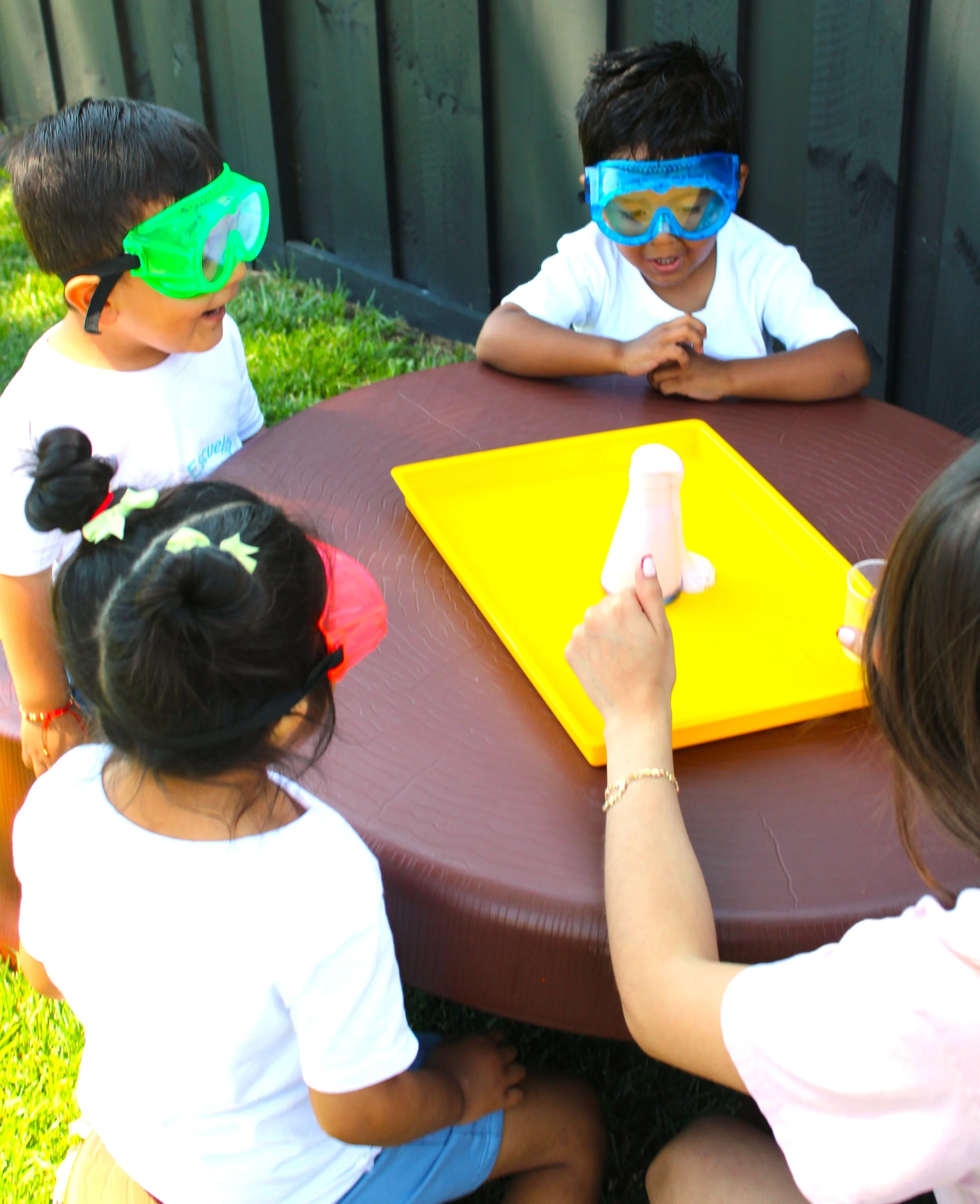 Two young boys wearing safety goggles, sitting around a brown outdoor table, engaged in a science experiment with a yellow tray and a model of a nose, outdoors in a grassy area.