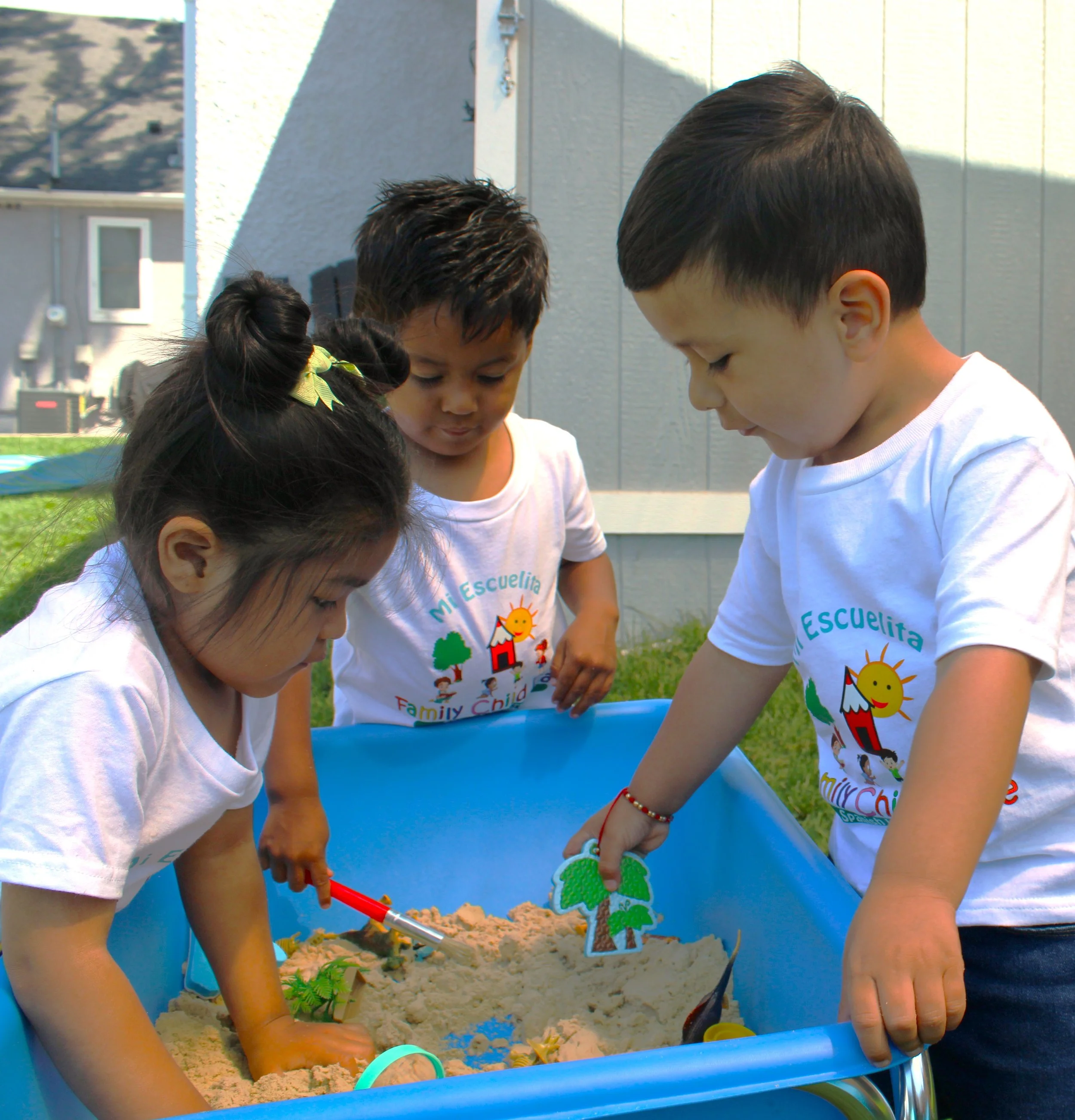 Children playing in a sandbox outdoors, wearing white shirts with a colorful logo and text, with some adults nearby.
